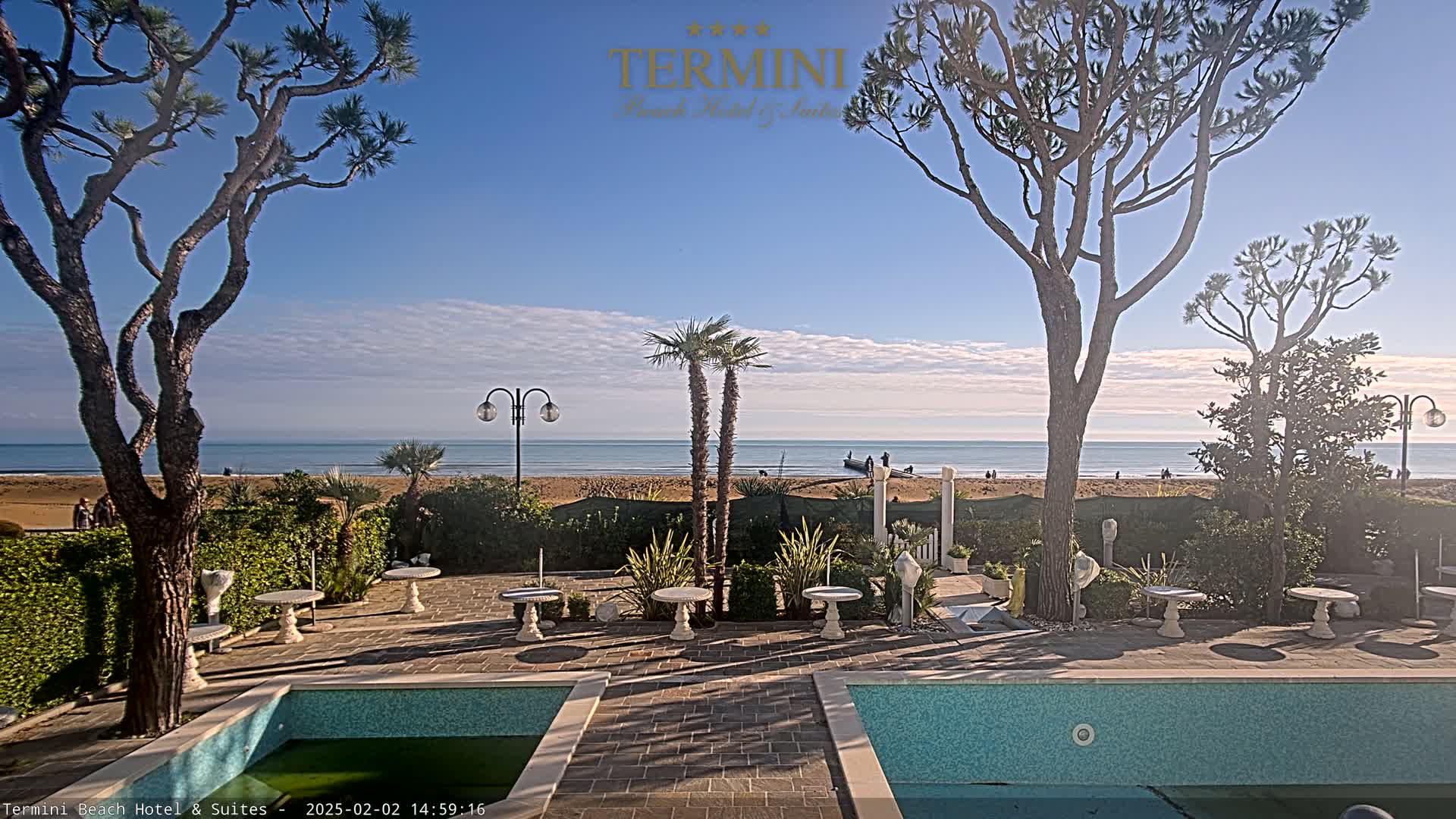 A sunny outdoor scene shows a patio with two small pools, landscaping, and tables overlooking a sandy beach and calm ocean.