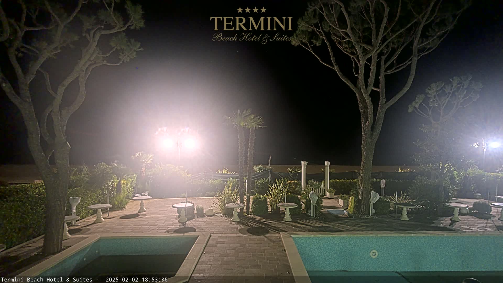 At night, a poolside patio with several small tables and trees is illuminated by bright lights, with a swimming pool visible in the foreground.