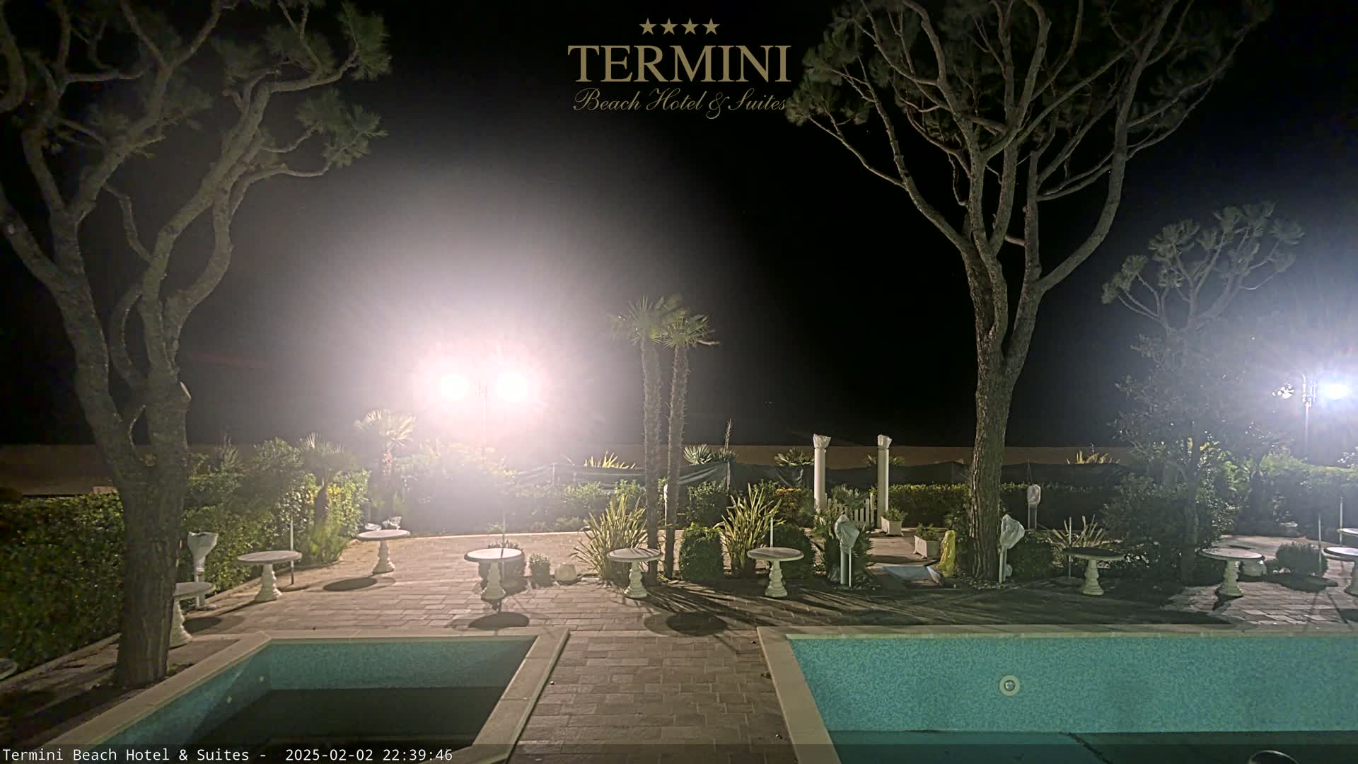 At night, a patio area with two swimming pools is illuminated by bright lights, surrounded by trees and landscaping.