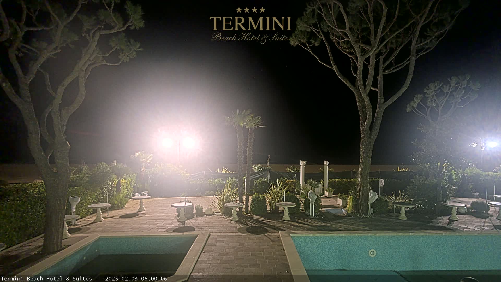 At night, a brightly lit patio area with two swimming pools is surrounded by trees and small plants.