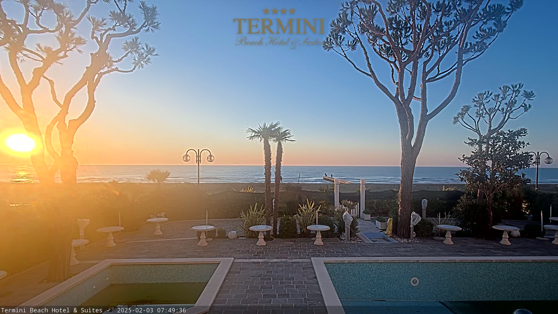 A sunrise over a calm ocean is viewed from a patio with two pools and several small tables, all surrounded by trees and landscaping.
