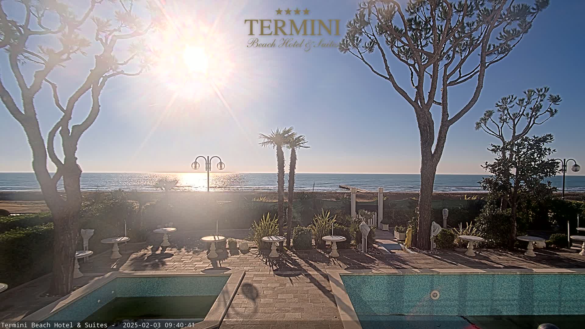 A sunny outdoor scene shows a patio area with small tables and a pool, overlooking a calm ocean under a clear sky.