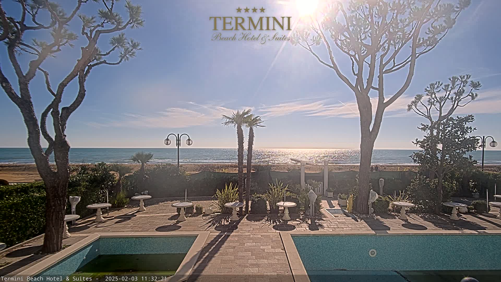 A sunny outdoor scene shows a patio area with small tables and a partially visible swimming pool, overlooking a calm ocean beach lined with trees.