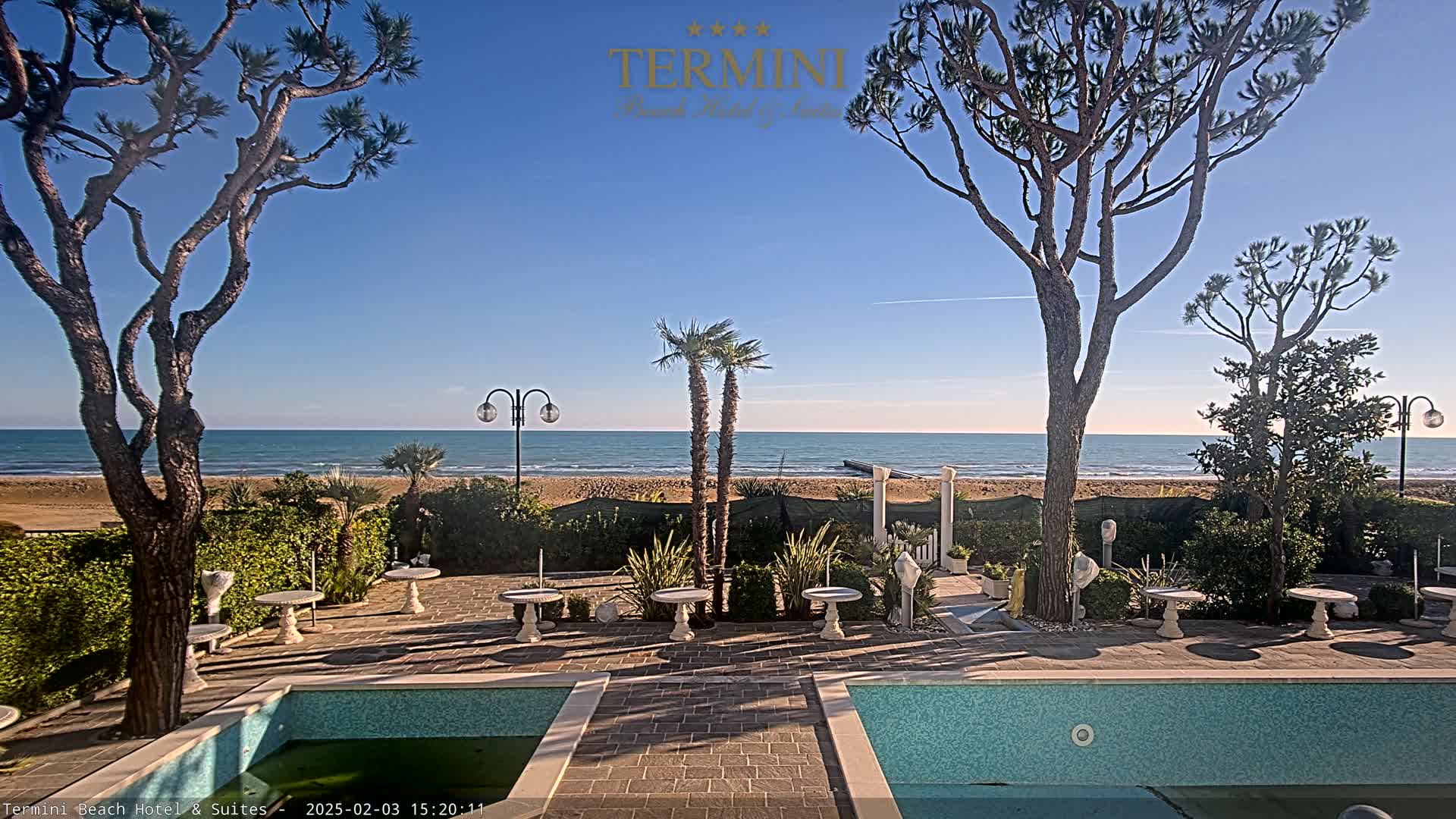 A patio area with two swimming pools overlooks a calm ocean on a sunny day.