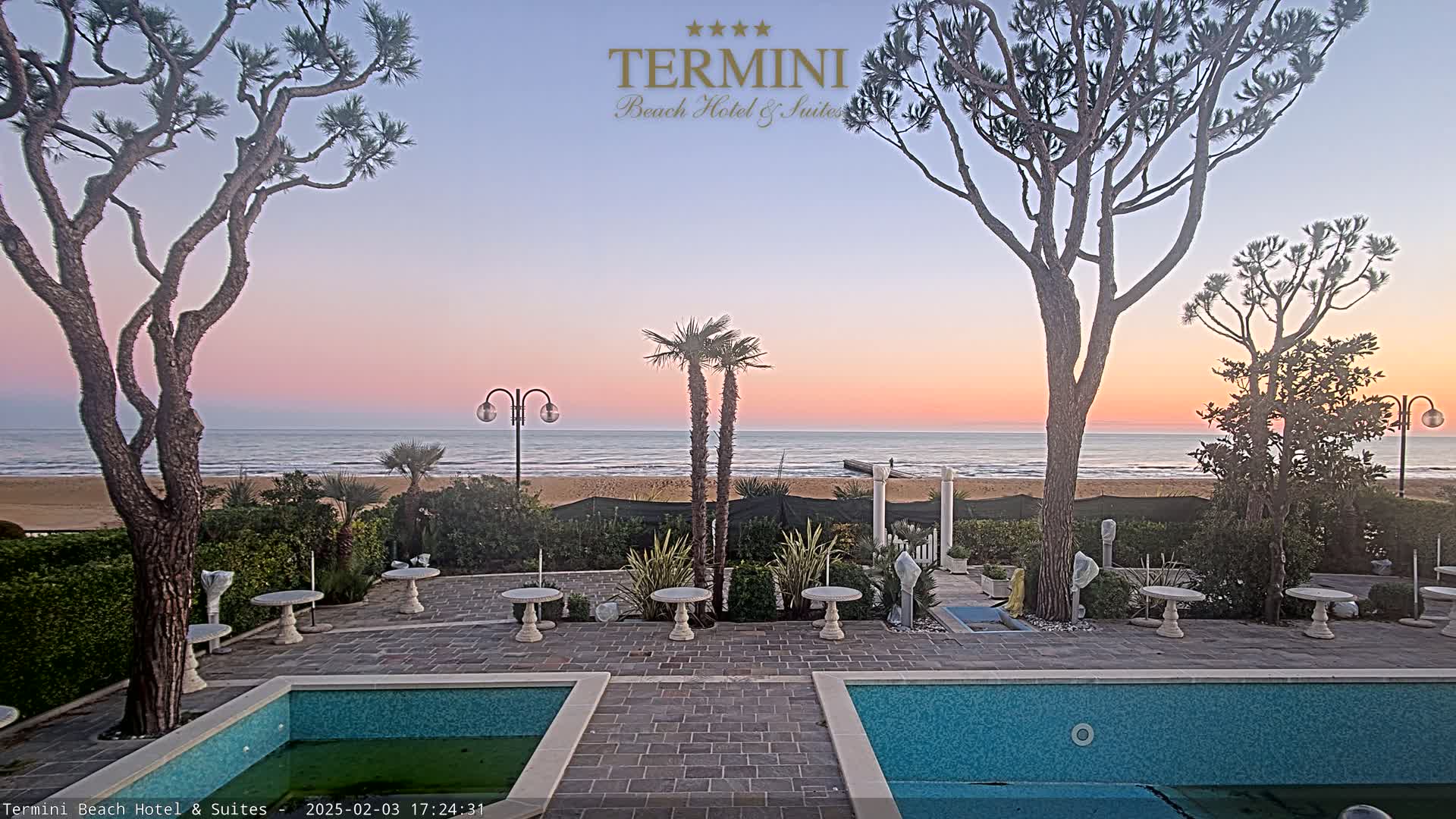 A patio with two pools overlooks a sandy beach and calm ocean under a sunset sky.