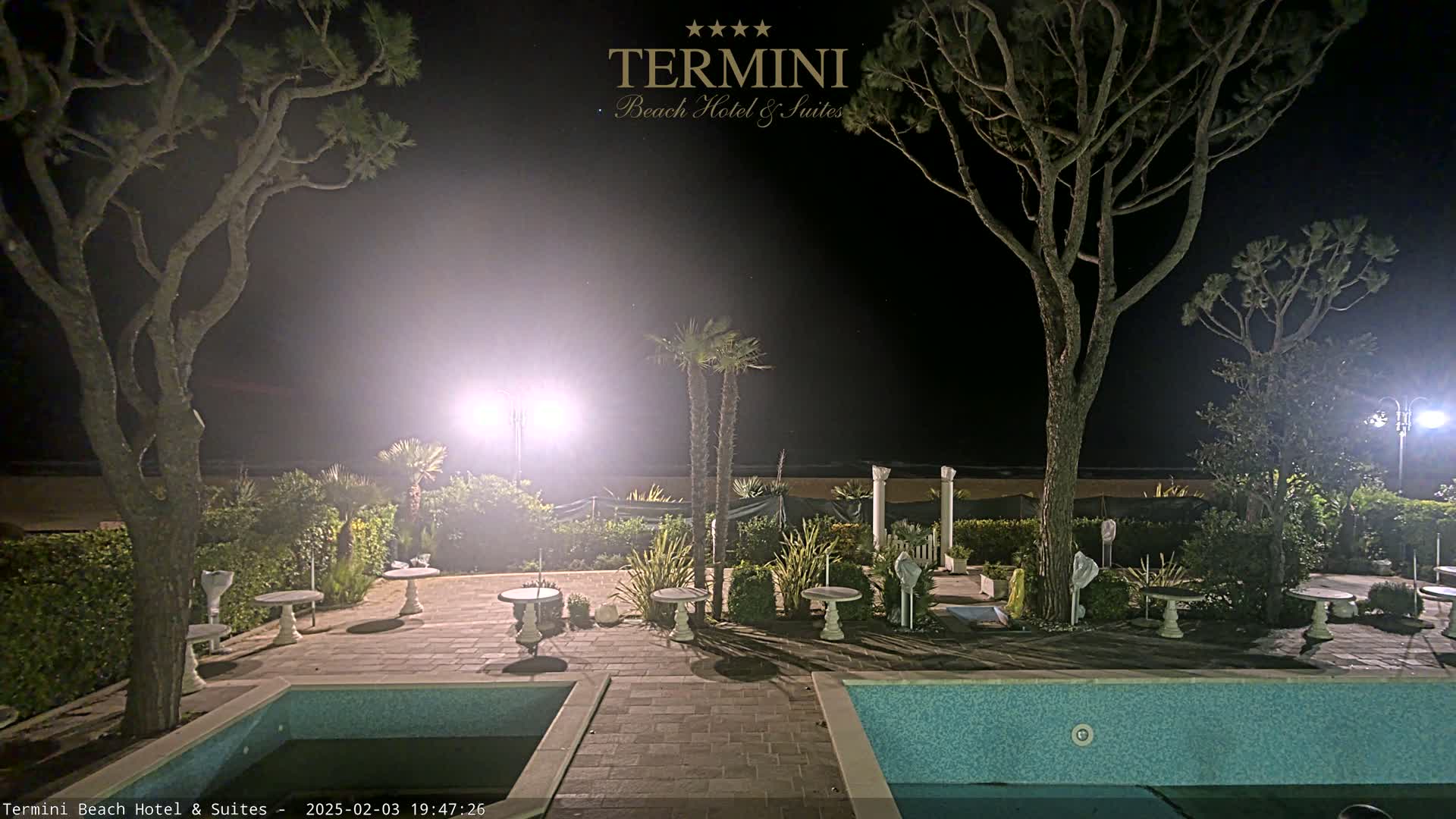 At night, an outdoor patio area with two swimming pools is illuminated, surrounded by trees and small tables.