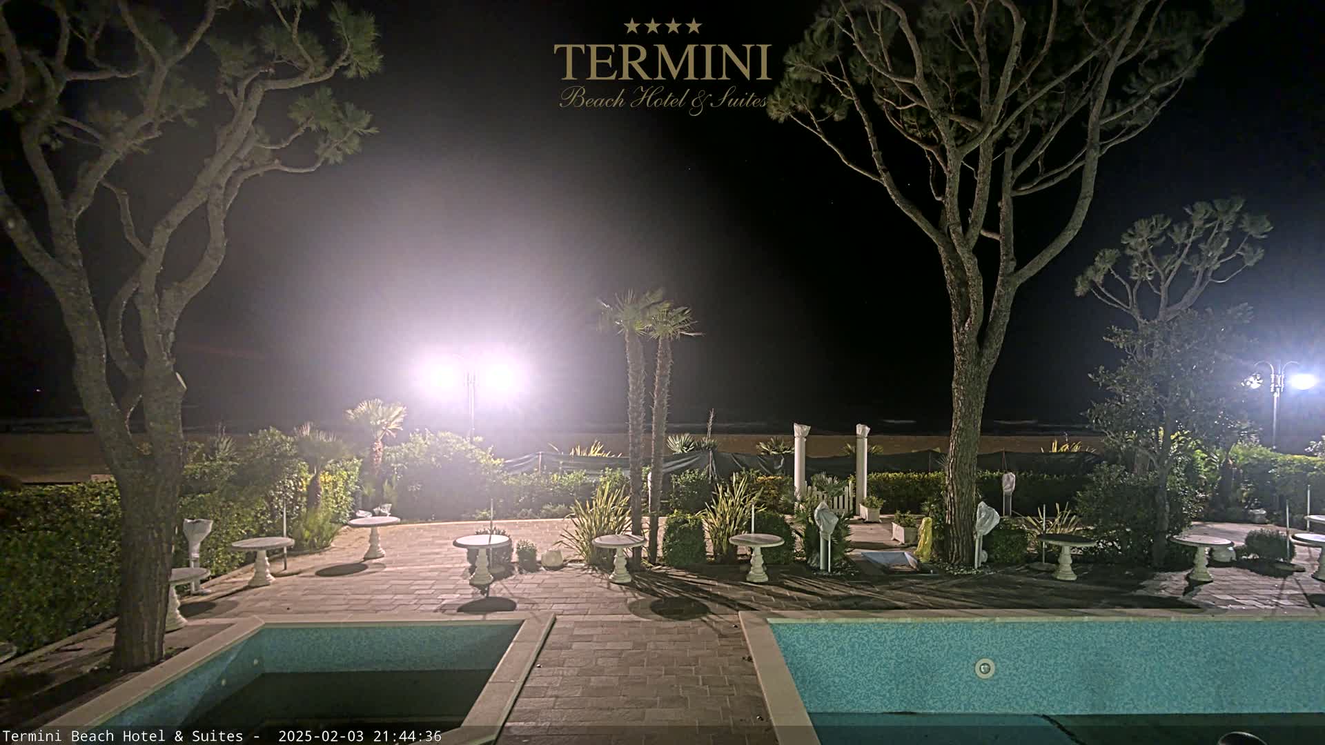 A nighttime view of a resort patio with two swimming pools, small tables, and palm trees, under hazy or foggy conditions.