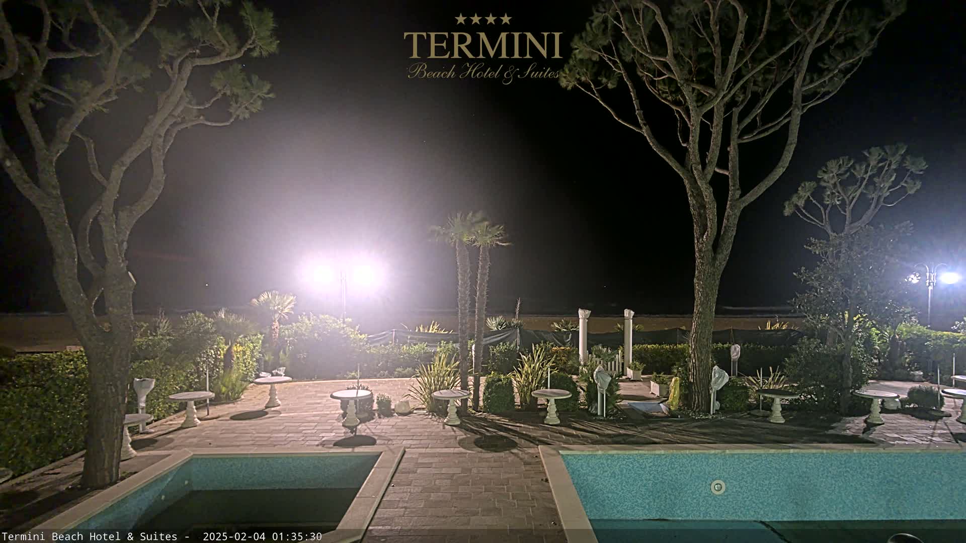 At night, a hazy outdoor scene shows two swimming pools and several small, round tables situated among palm trees and pine trees under bright lights.