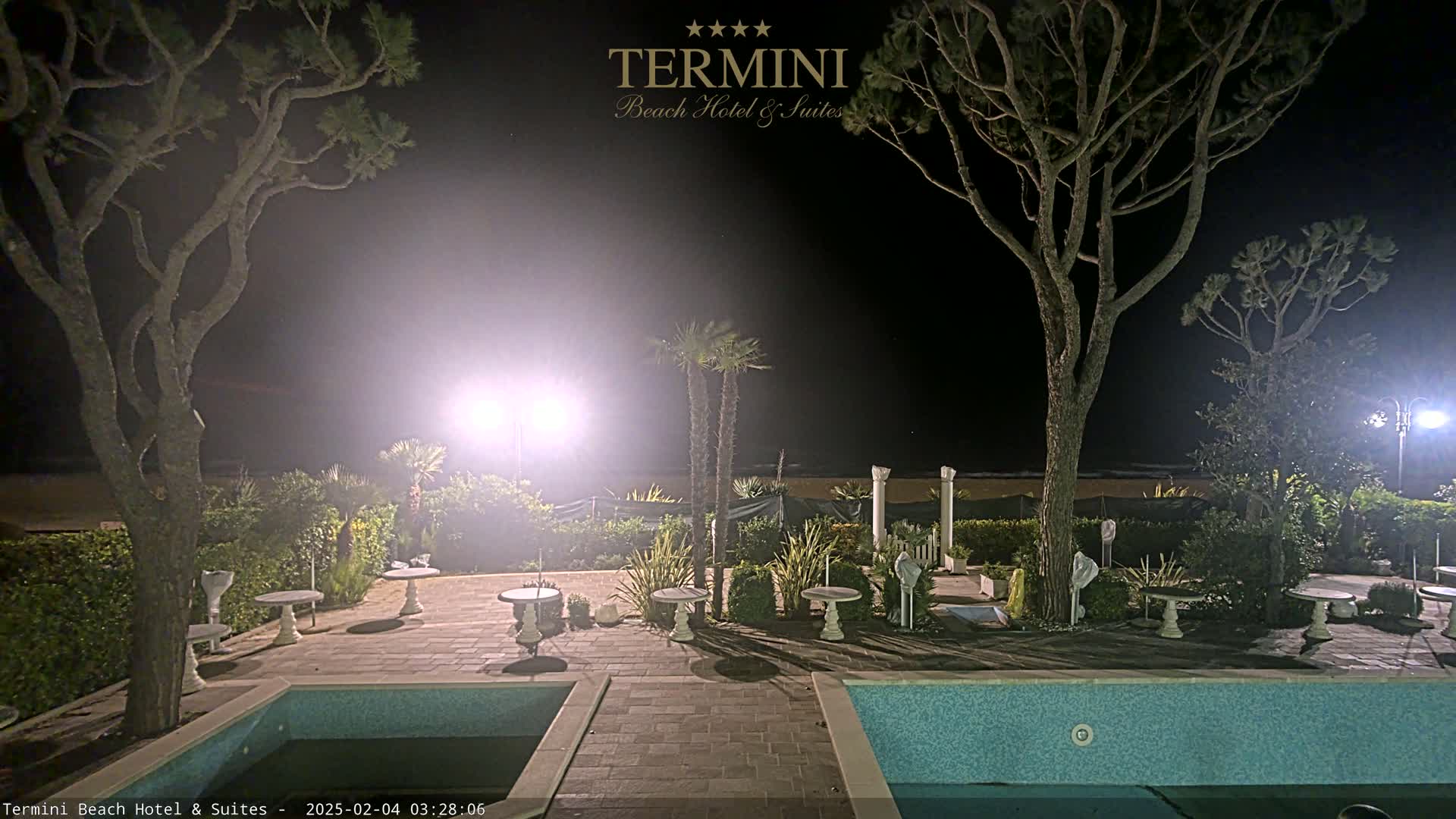 A nighttime view of a patio area with two pools, small tables, and trees, illuminated by artificial lights, overlooking a beach.