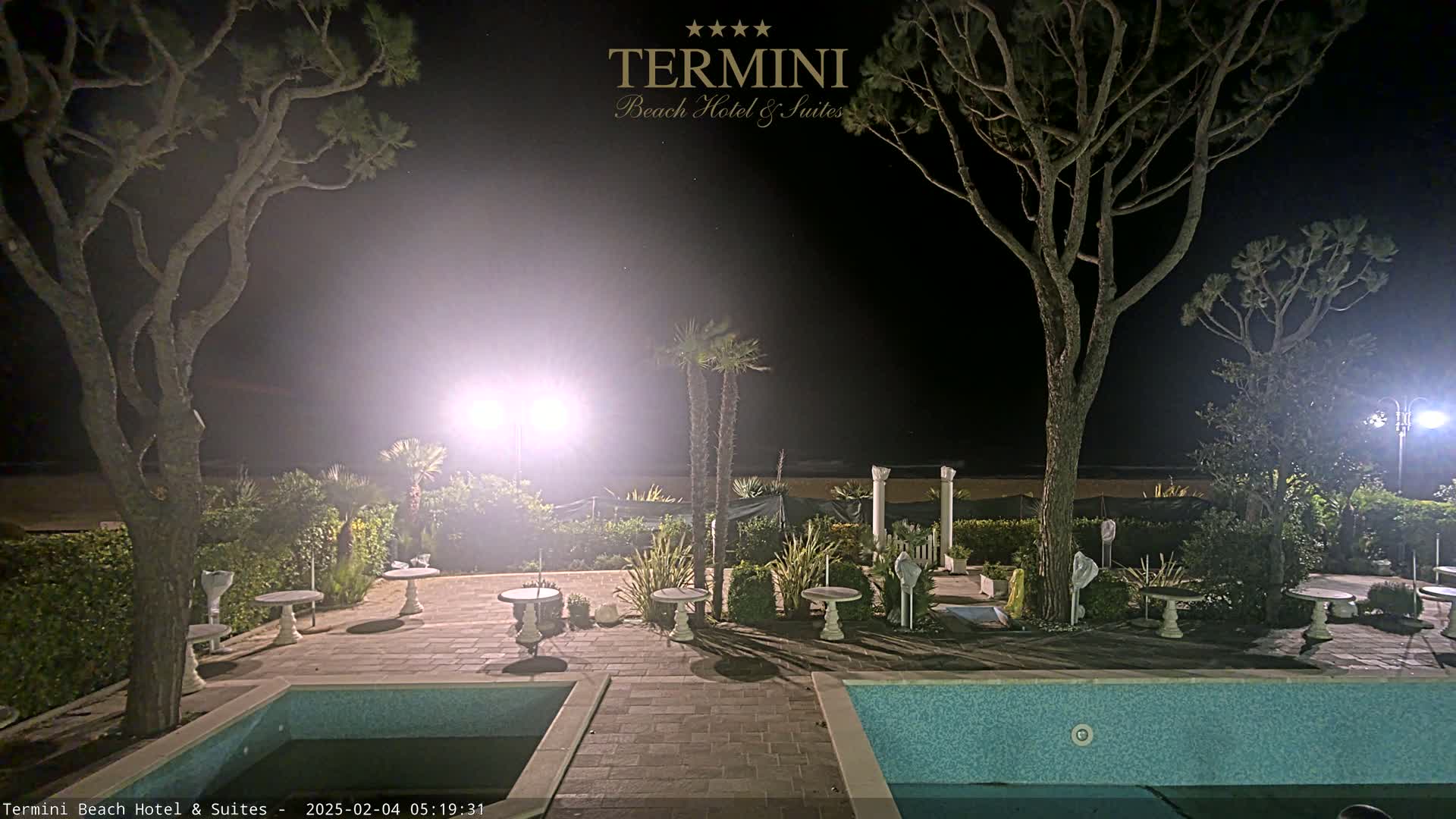 At night, a hotel pool area with palm trees and outdoor furniture is illuminated by bright lights.
