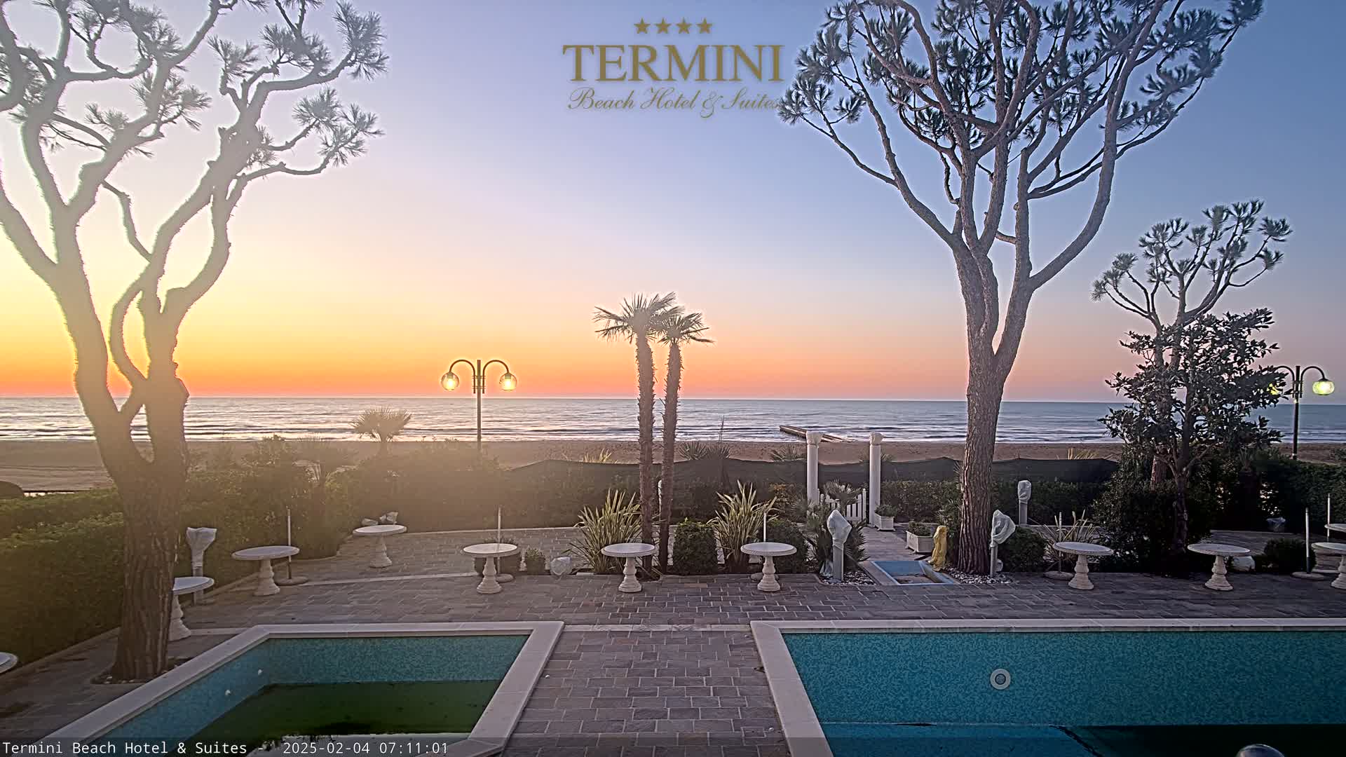 A sunrise over a calm ocean is viewed from a patio area with two swimming pools,  trees, and small tables and chairs.