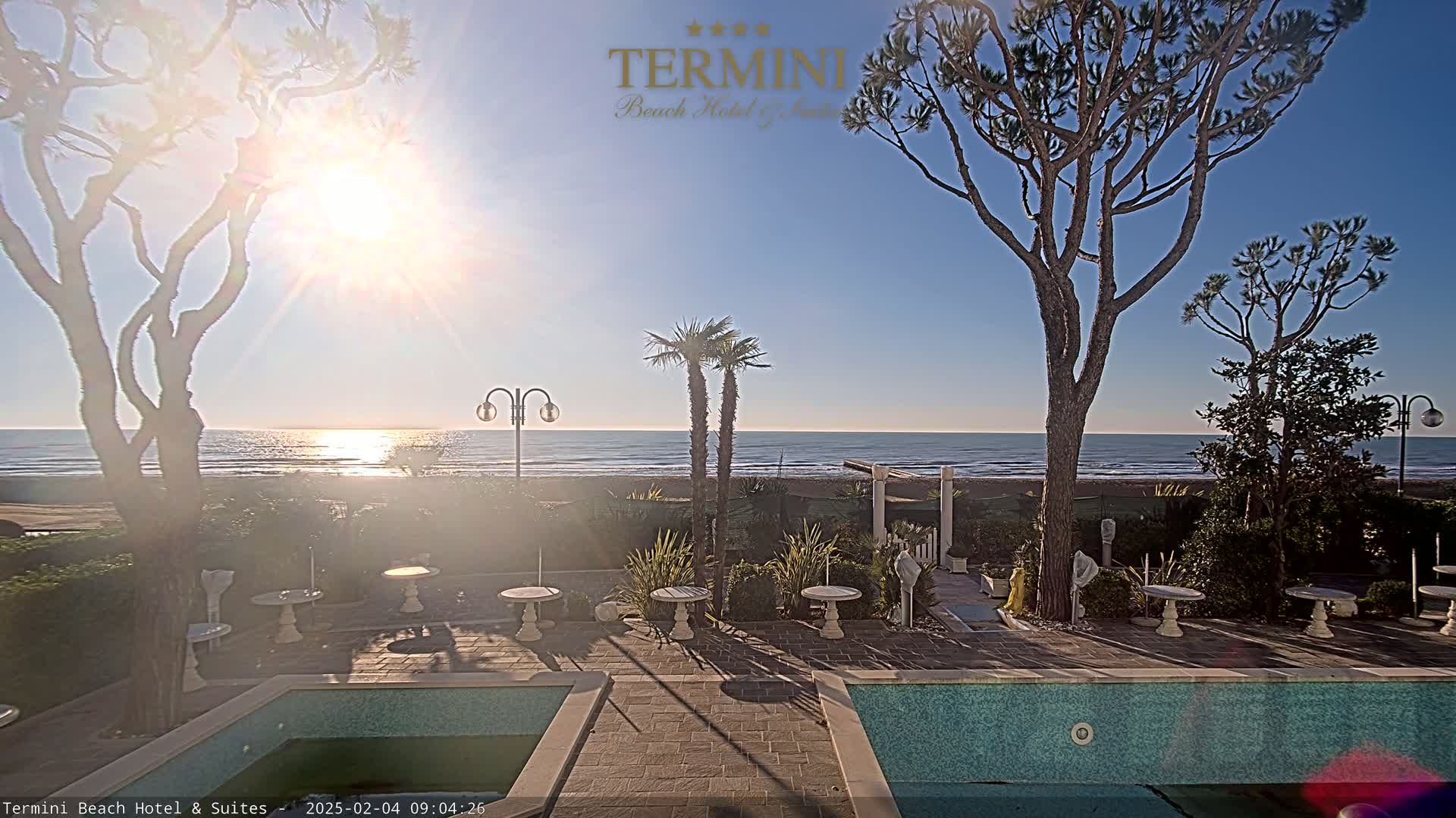A sunny outdoor scene shows a patio area with small tables, landscaping, and two pools overlooking a calm ocean beach.
