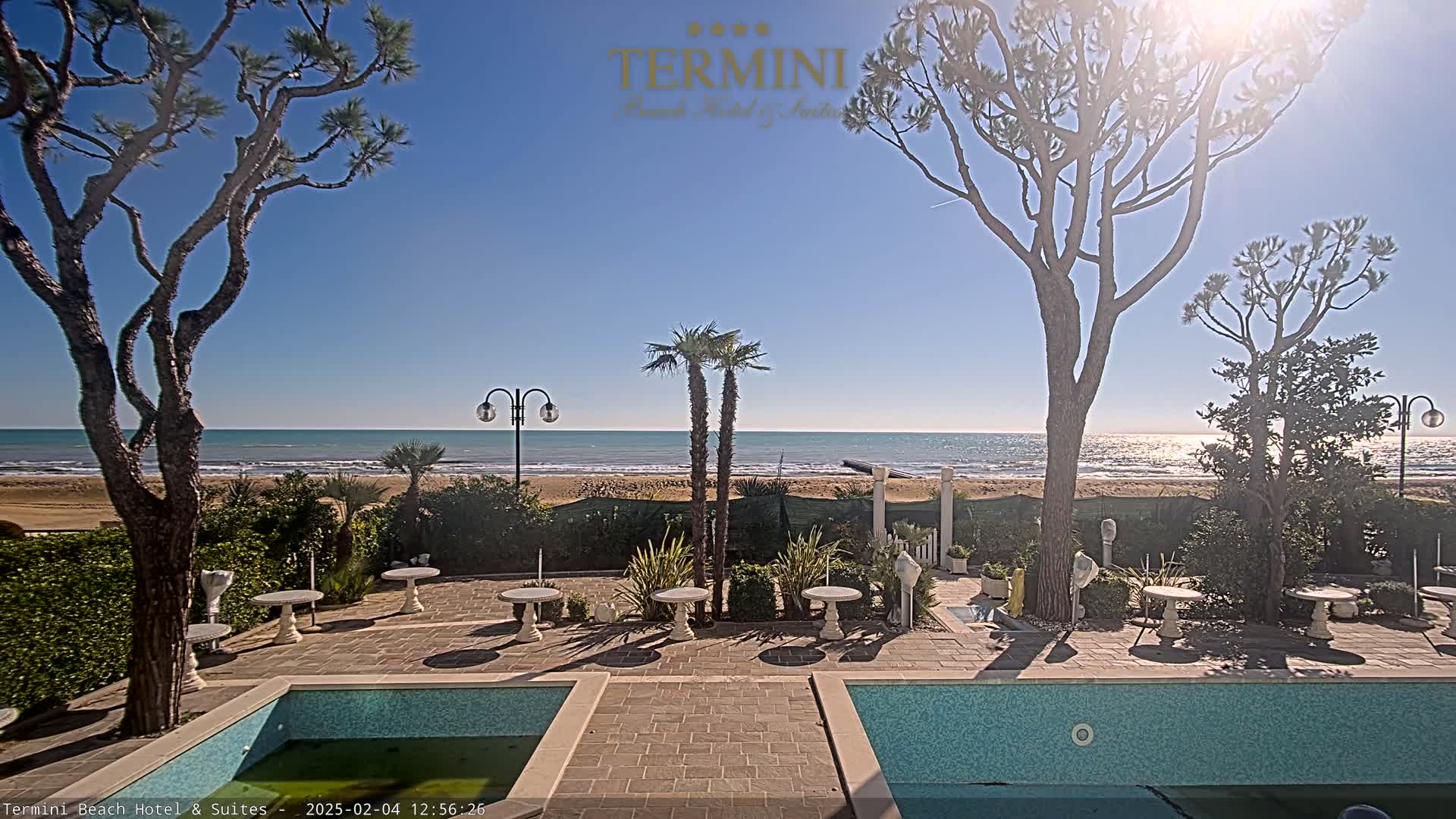 A sunny outdoor scene shows a patio area with a swimming pool and small tables, situated between tall trees and a sandy beach with a calm ocean under a clear blue sky.
