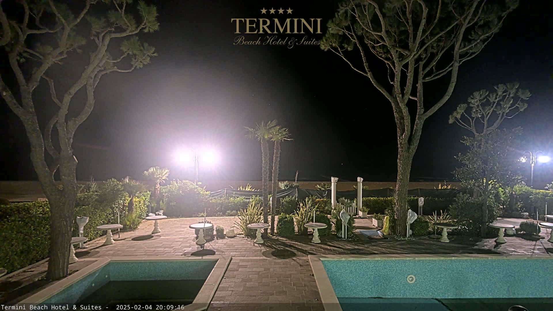 At night, an outdoor patio area with two swimming pools is illuminated, surrounded by trees and small tables.