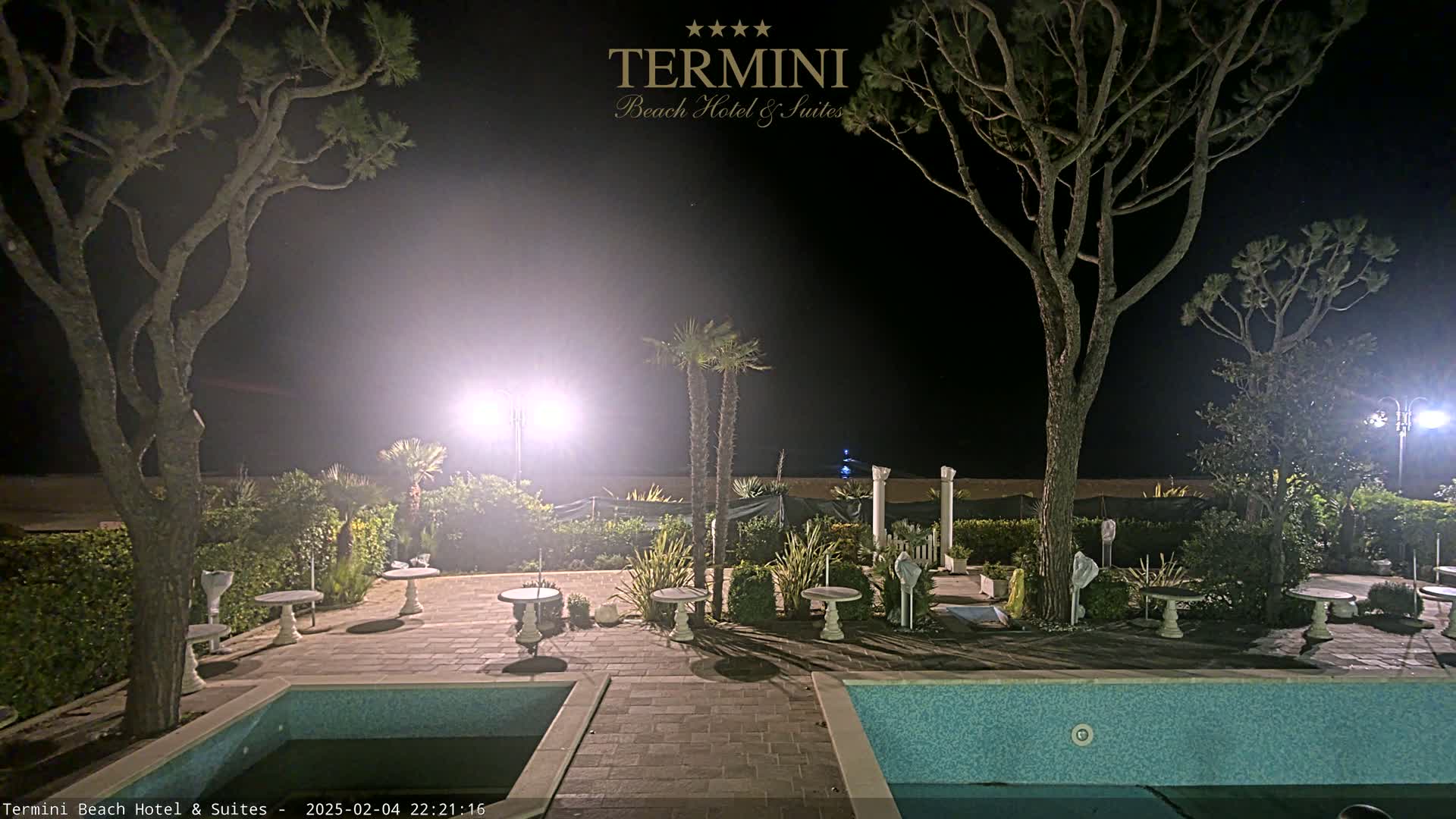 At night, a pool area with several small tables and two pools is illuminated by artificial lights, with tall trees and palm trees surrounding it, near a beach.