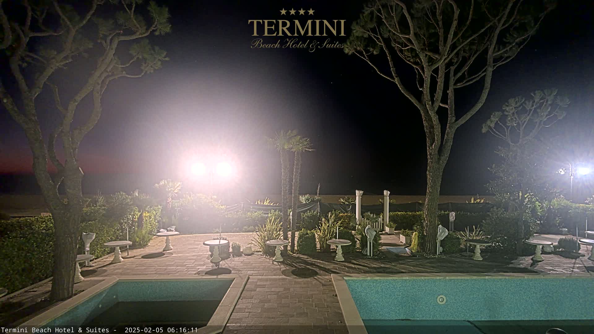 A nighttime view of a pool area with patio furniture, landscaping, and trees under bright lights, near a beach.