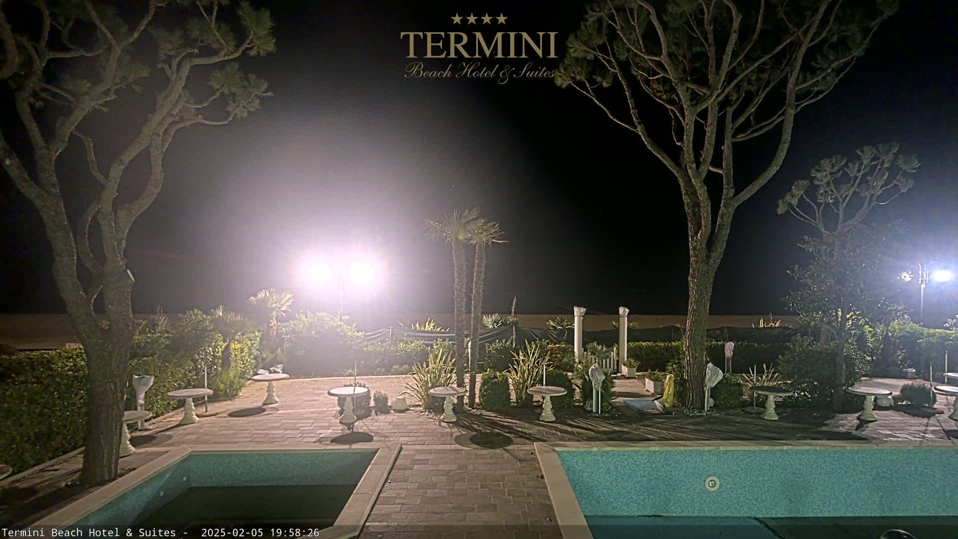 A nighttime view of a patio area with two swimming pools, several small tables, and palm trees illuminated by bright lights.