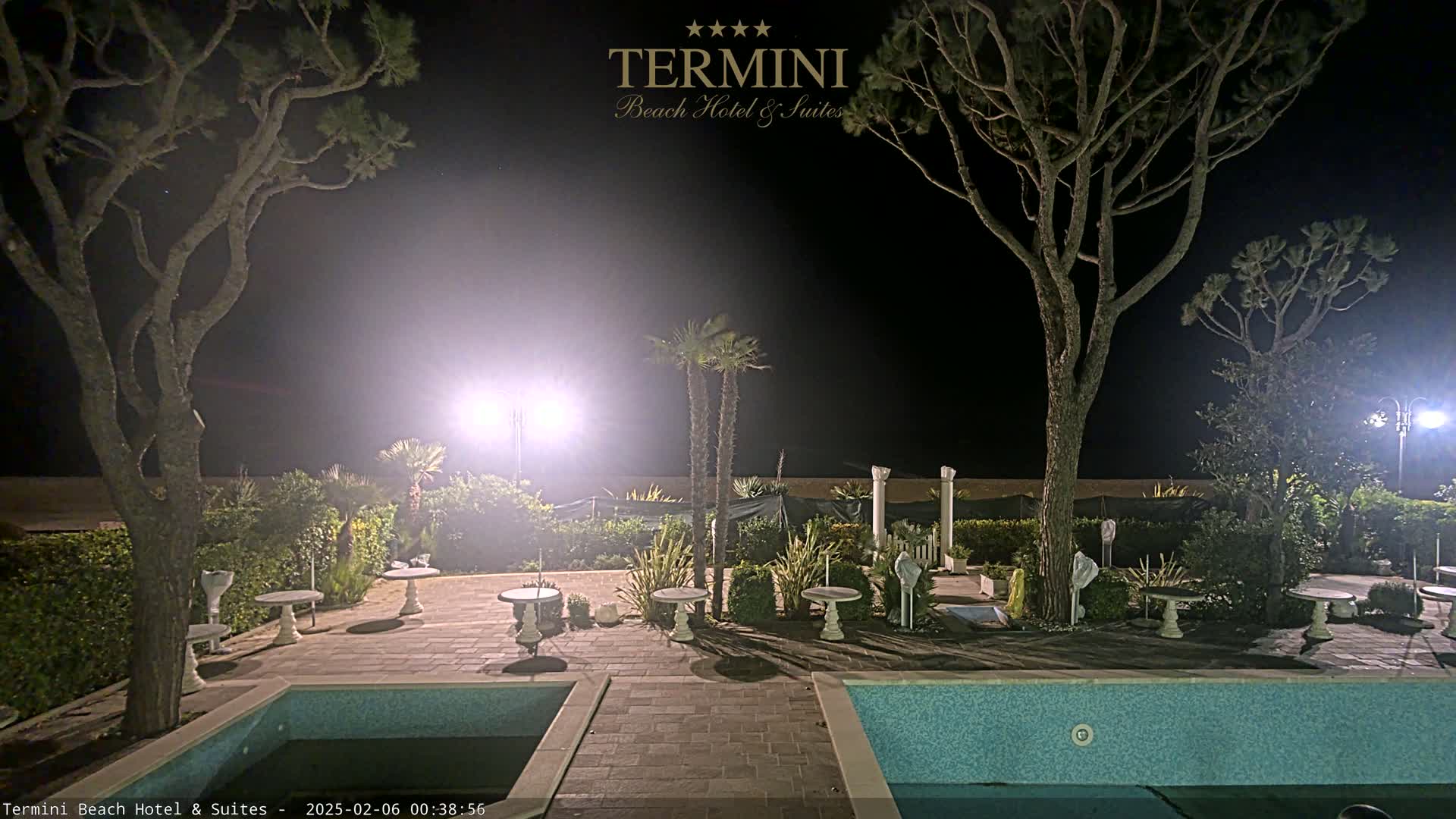 At night, a resort patio with two swimming pools is illuminated by bright lights, surrounded by trees and landscaping.