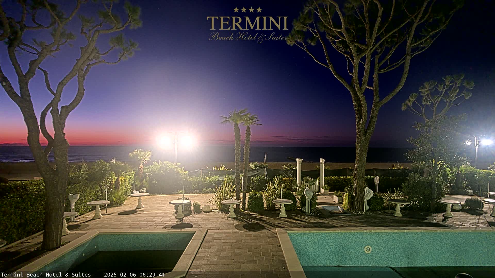 A twilight scene shows a resort patio with a swimming pool, small tables, palm trees, and two tall, slender trees, all overlooking a calm ocean under a purplish-pink sky.