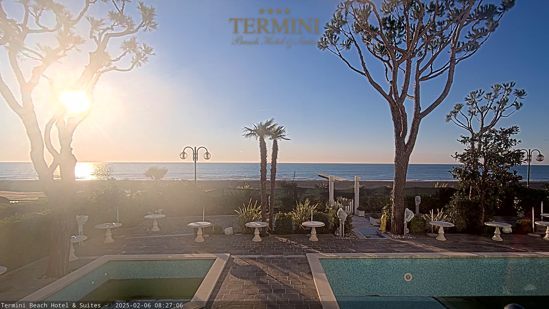 A sunny seaside scene features two swimming pools, patio furniture, and palm and pine trees overlooking a calm ocean under a clear sky.