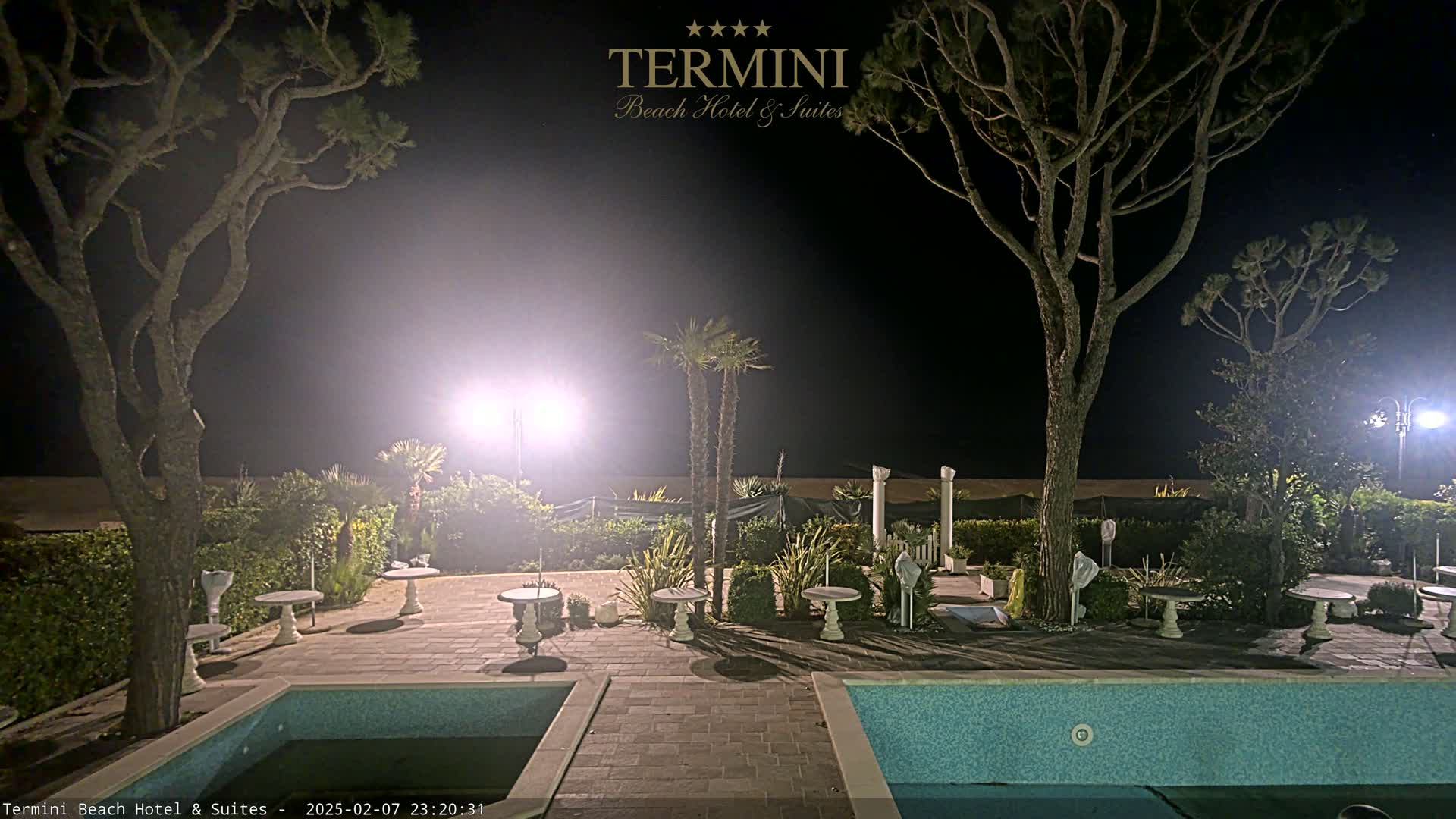 A nighttime view of a resort courtyard with two swimming pools, several small tables, and palm trees illuminated by bright lights.