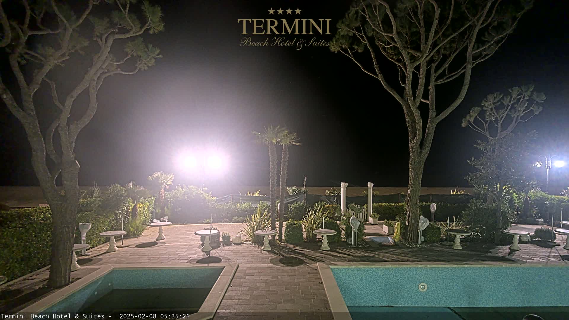 A nighttime view of a hotel patio area with two swimming pools, several small tables and chairs, palm trees, and tall pine trees, illuminated by bright lights.