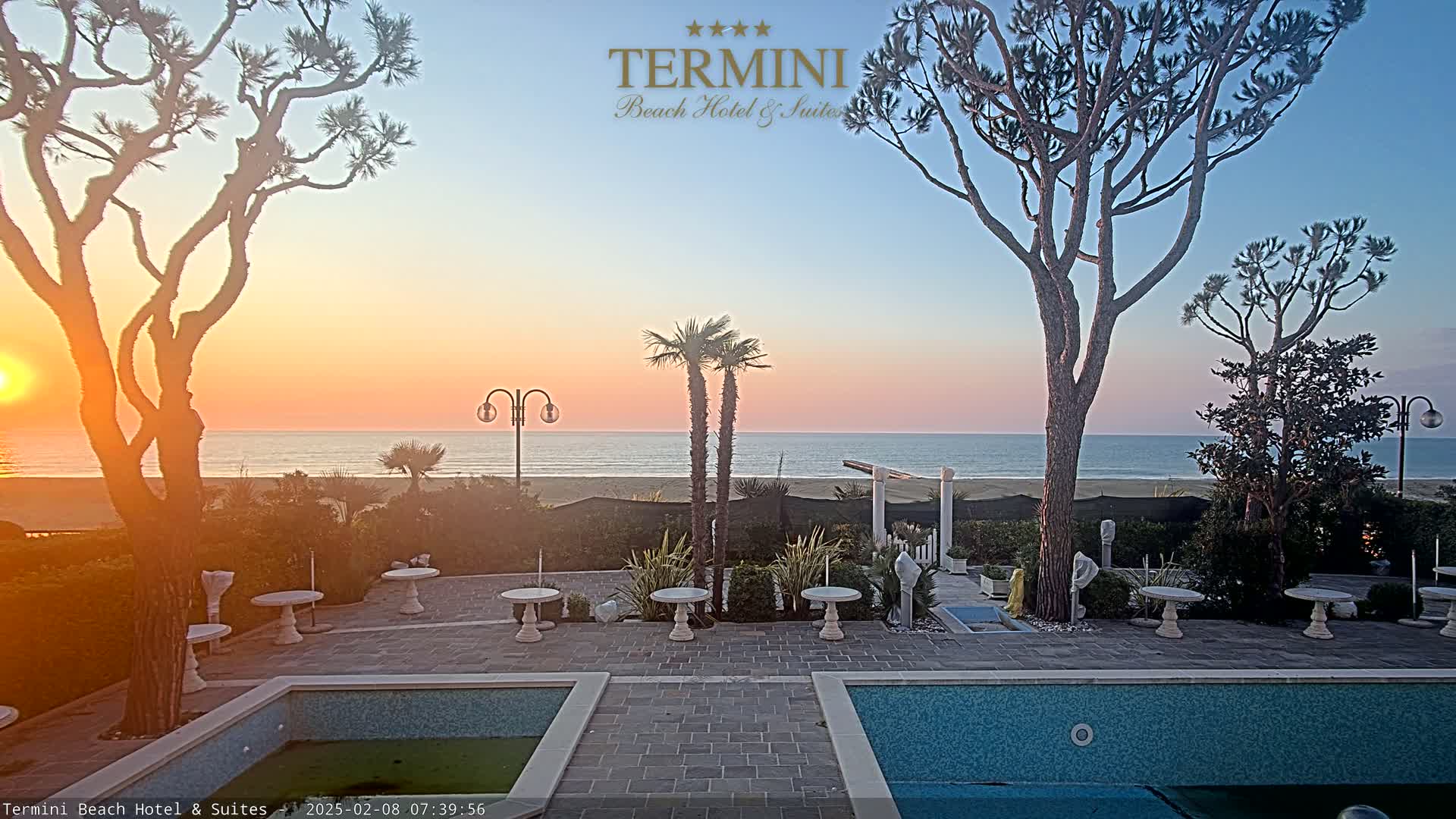 A calm ocean at sunset is viewed from a patio with two pools, several small tables, and trees.