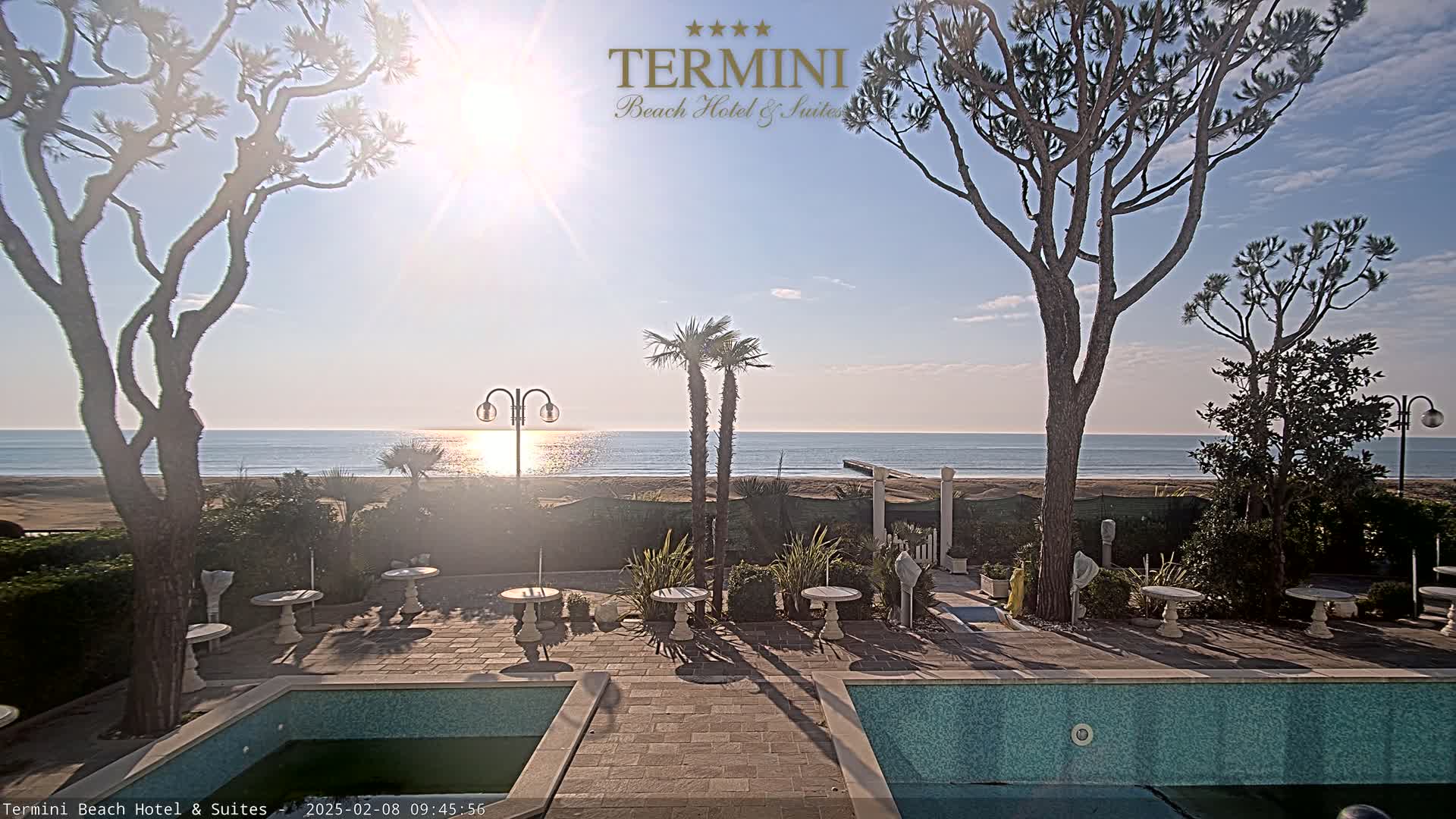 A sunny day at a beachfront hotel shows a calm ocean, a sandy beach, palm trees, and an outdoor pool area with patio furniture.