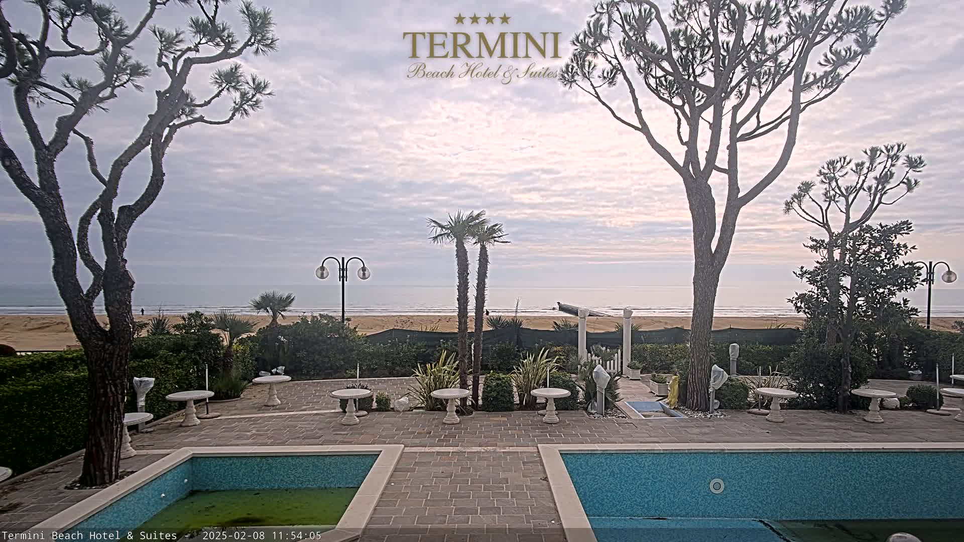A patio with two small swimming pools overlooks a sandy beach and calm ocean under a cloudy sky.