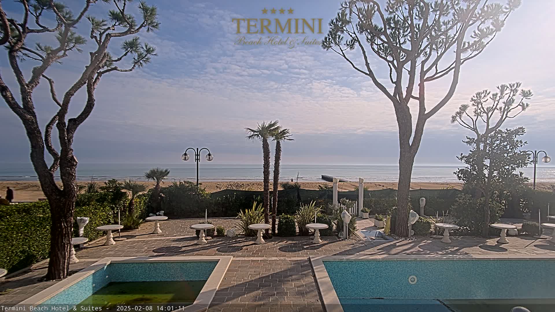 A patio with two small pools overlooks a calm ocean beach under a mostly sunny sky.