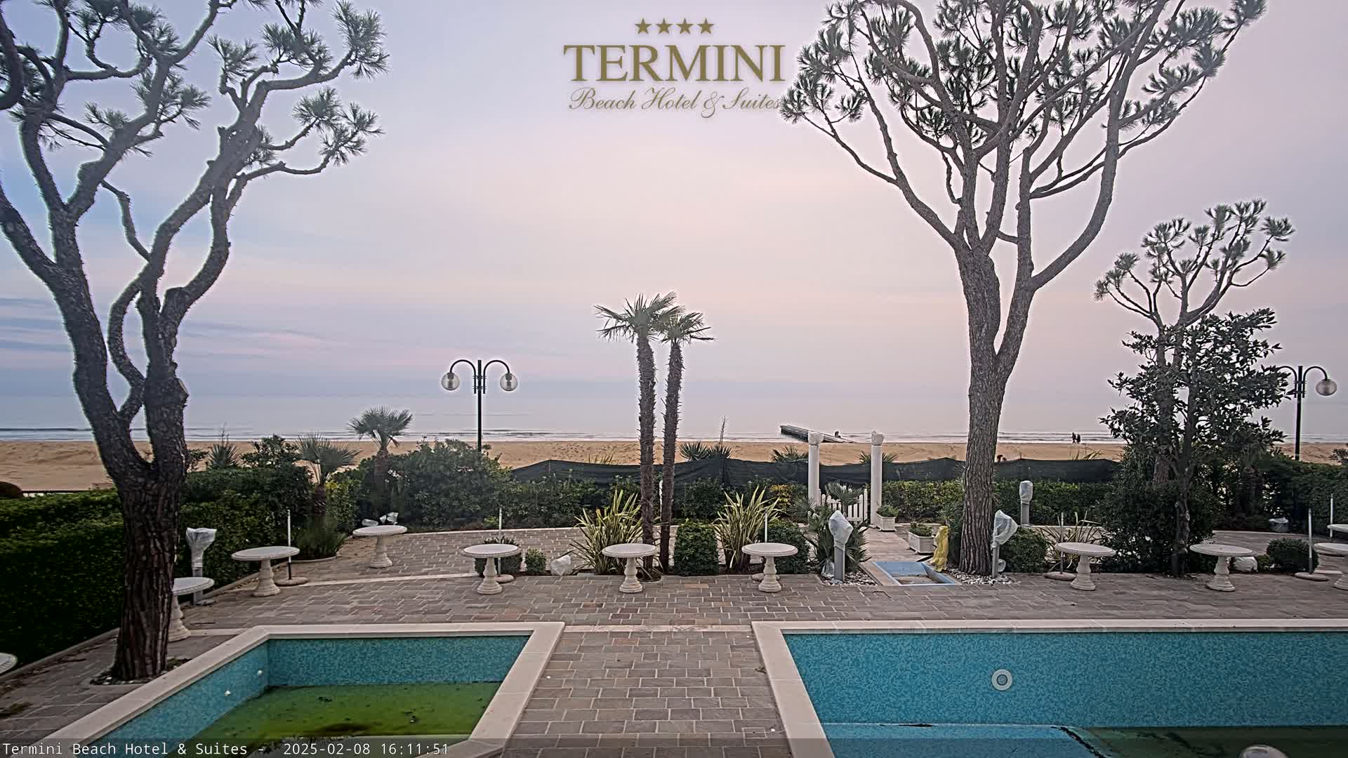 An outdoor patio area with two small pools overlooks a calm ocean beach under a pale sky.