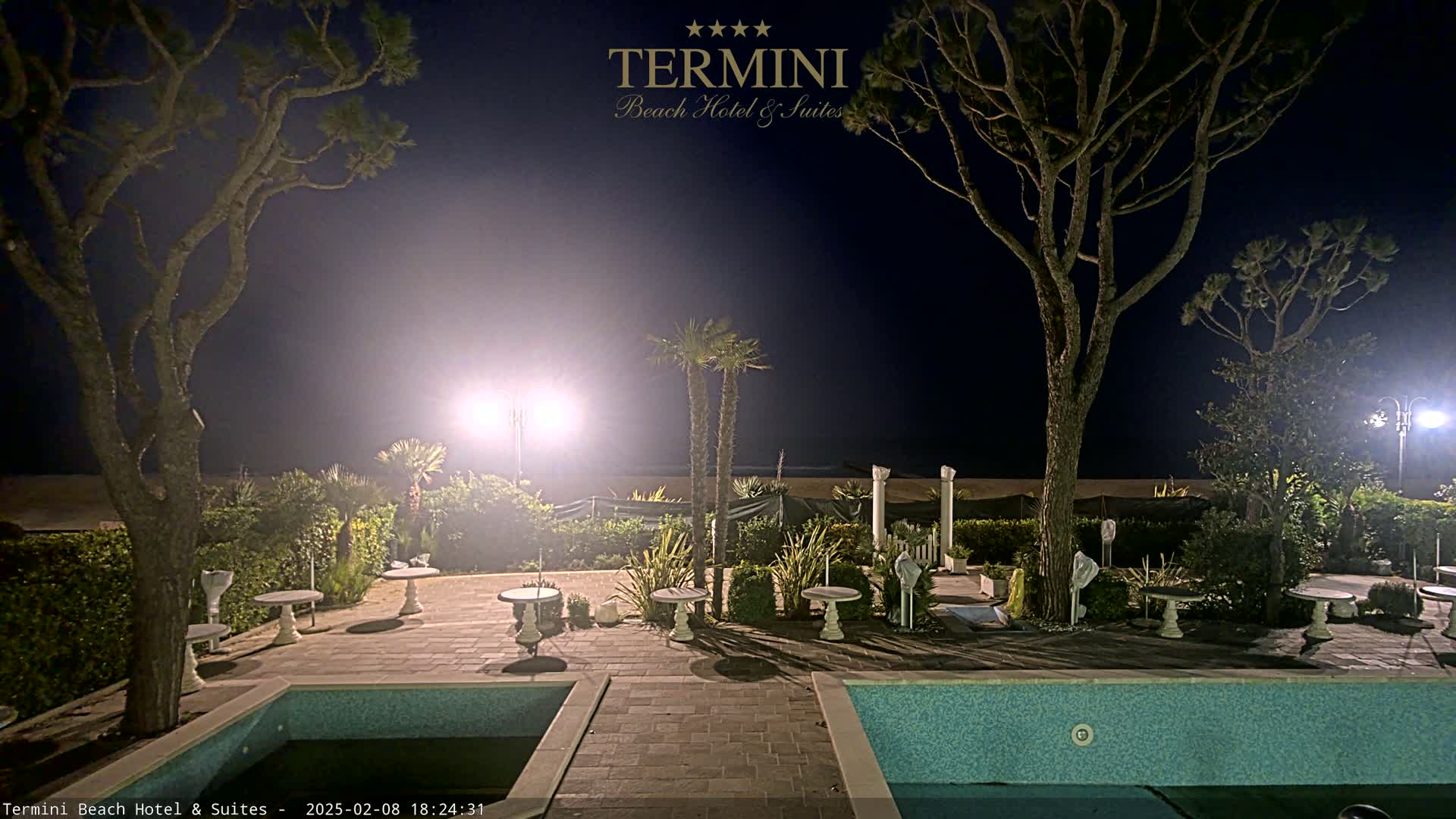 At night, a hazy, dimly lit outdoor patio area with two swimming pools, several small tables, and landscaping features under tall trees is seen next to a beach.
