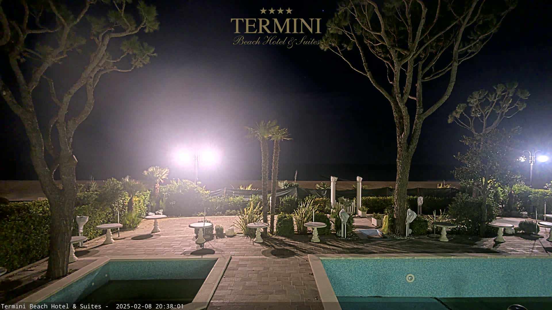 At night, a courtyard with two swimming pools is illuminated, surrounded by trees, landscaping, and small tables.