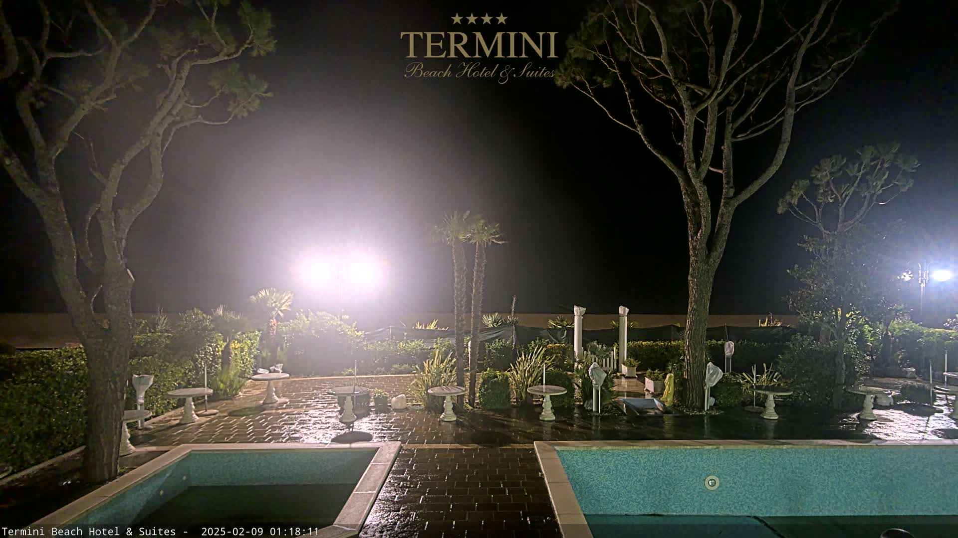 At night, a patio area with two swimming pools is illuminated, surrounded by trees and landscaping.
