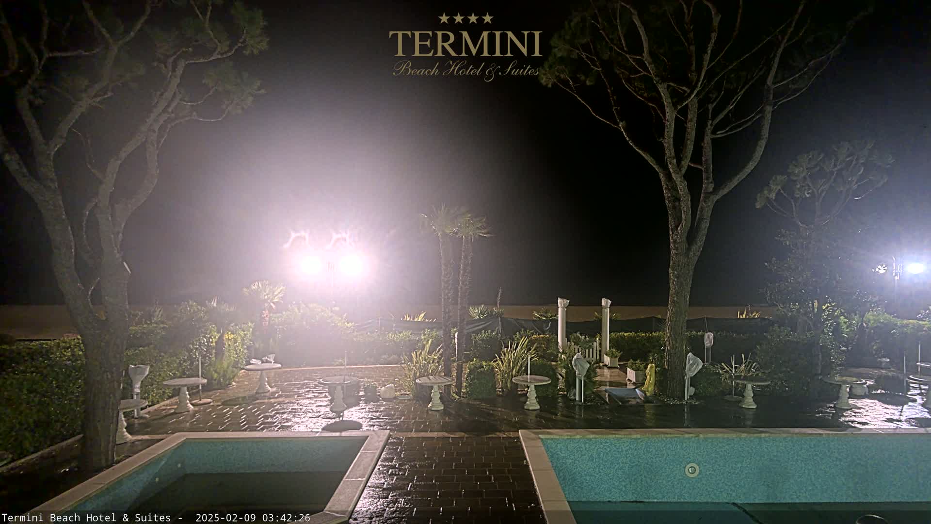 At night, under hazy, bright lights, an outdoor patio area with two swimming pools and several small tables is surrounded by trees and landscaping.