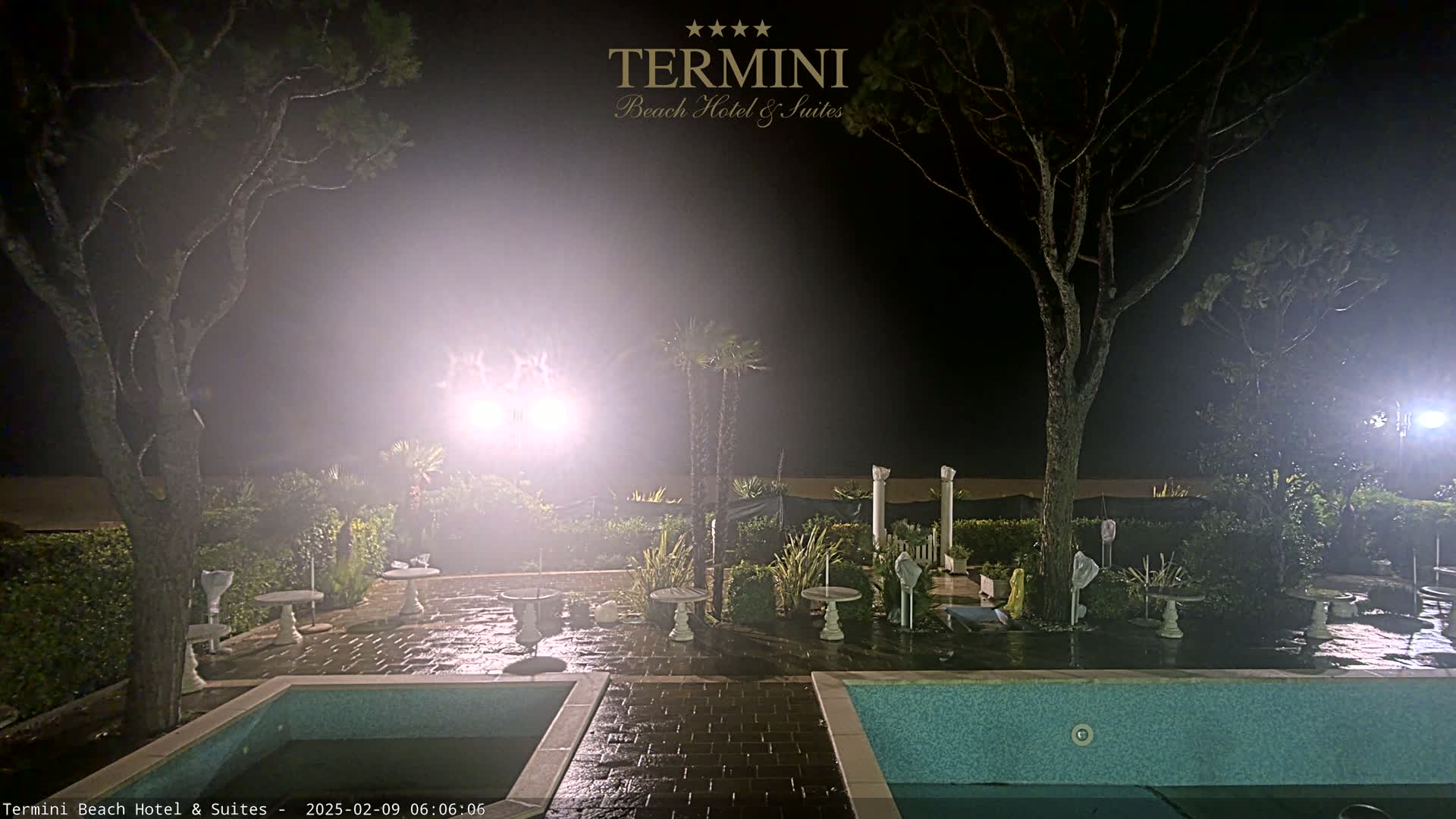 A foggy night scene shows an outdoor patio area with two pools, several small tables and chairs, and trees surrounding it, all illuminated by bright lights.