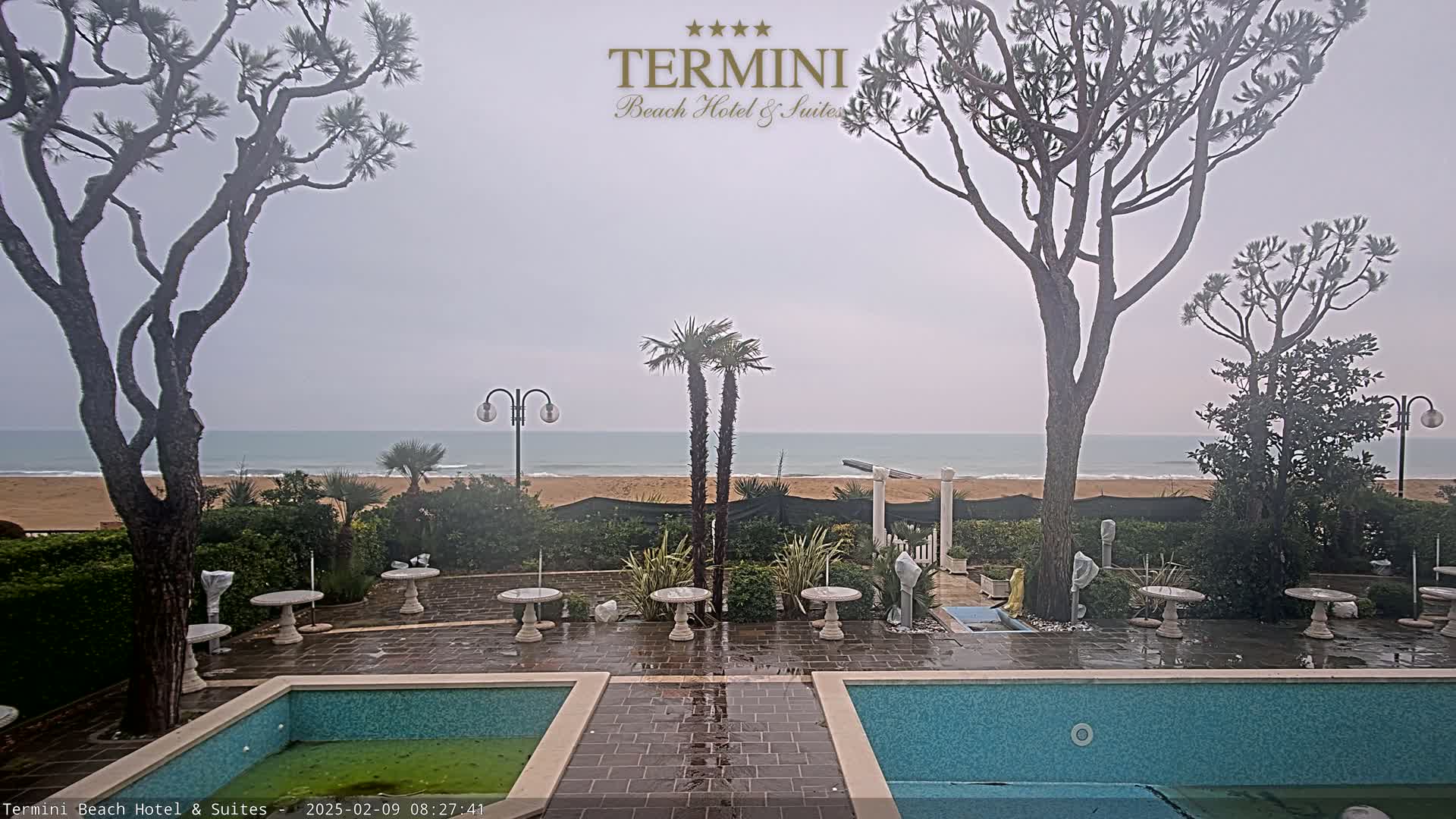 A wet patio with two pools overlooks a beach and ocean under an overcast sky.