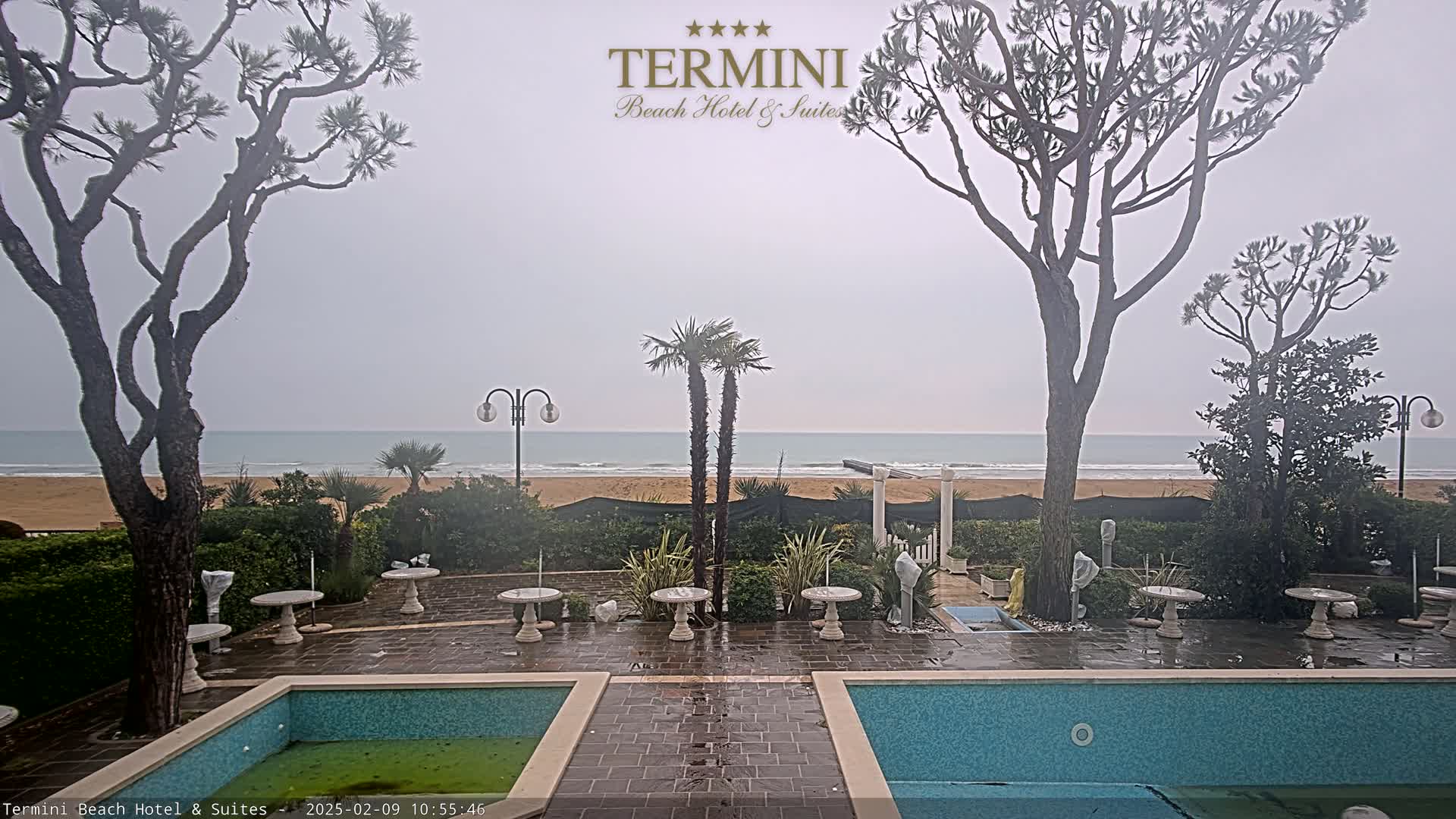 A view of a sandy beach and calm ocean on an overcast day, seen from a patio with two pools and several small tables, surrounded by trees and landscaping.