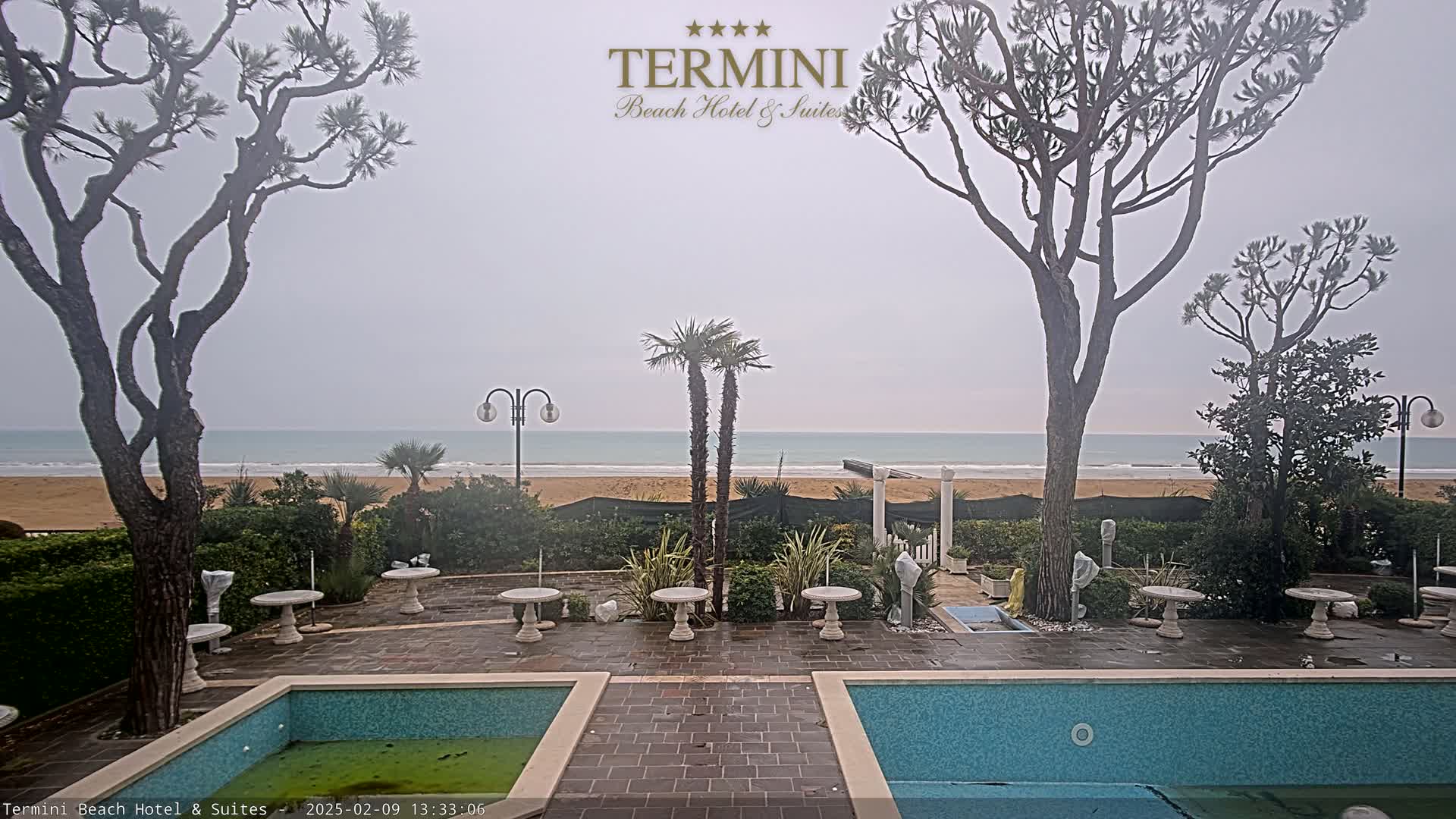 A patio with two pools overlooks a sandy beach and calm ocean under an overcast sky.