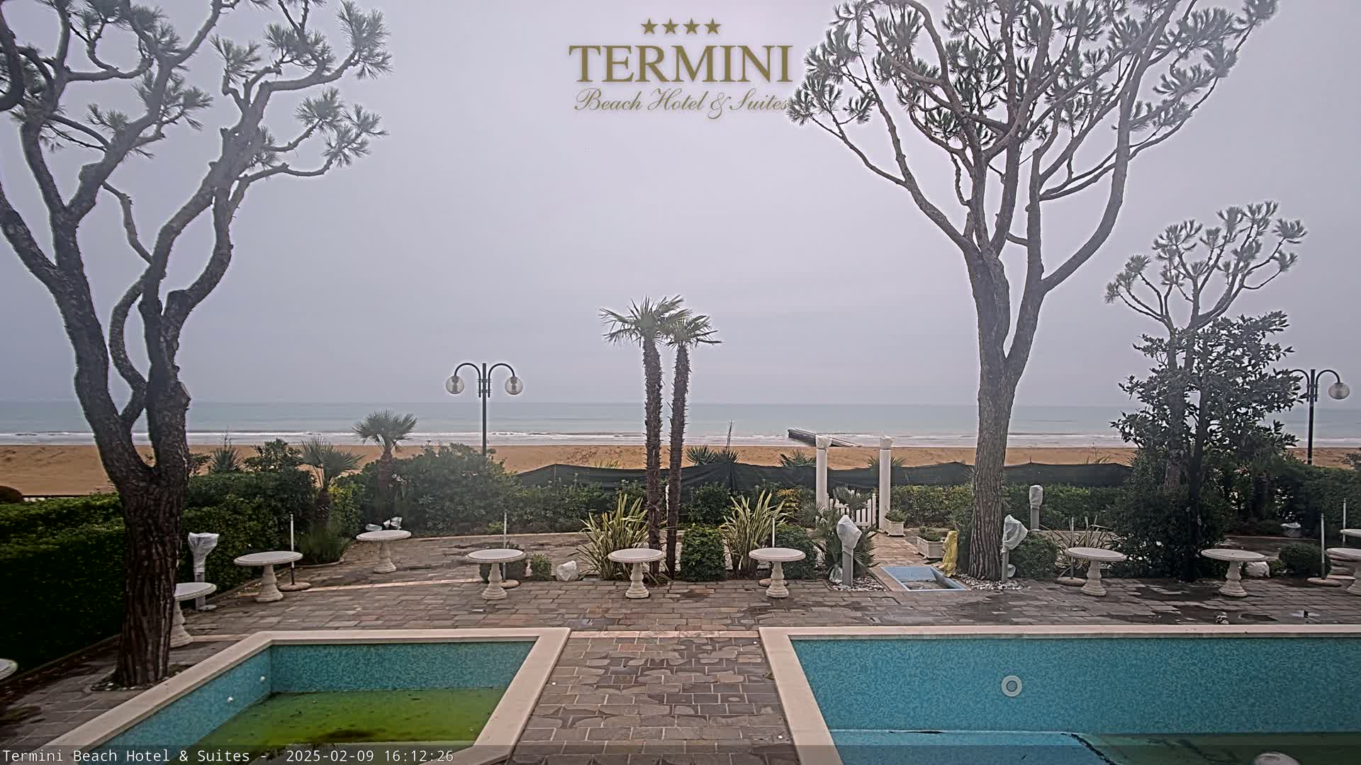 A view of a hotel courtyard with two swimming pools, overlooking a sandy beach and a calm, overcast ocean.