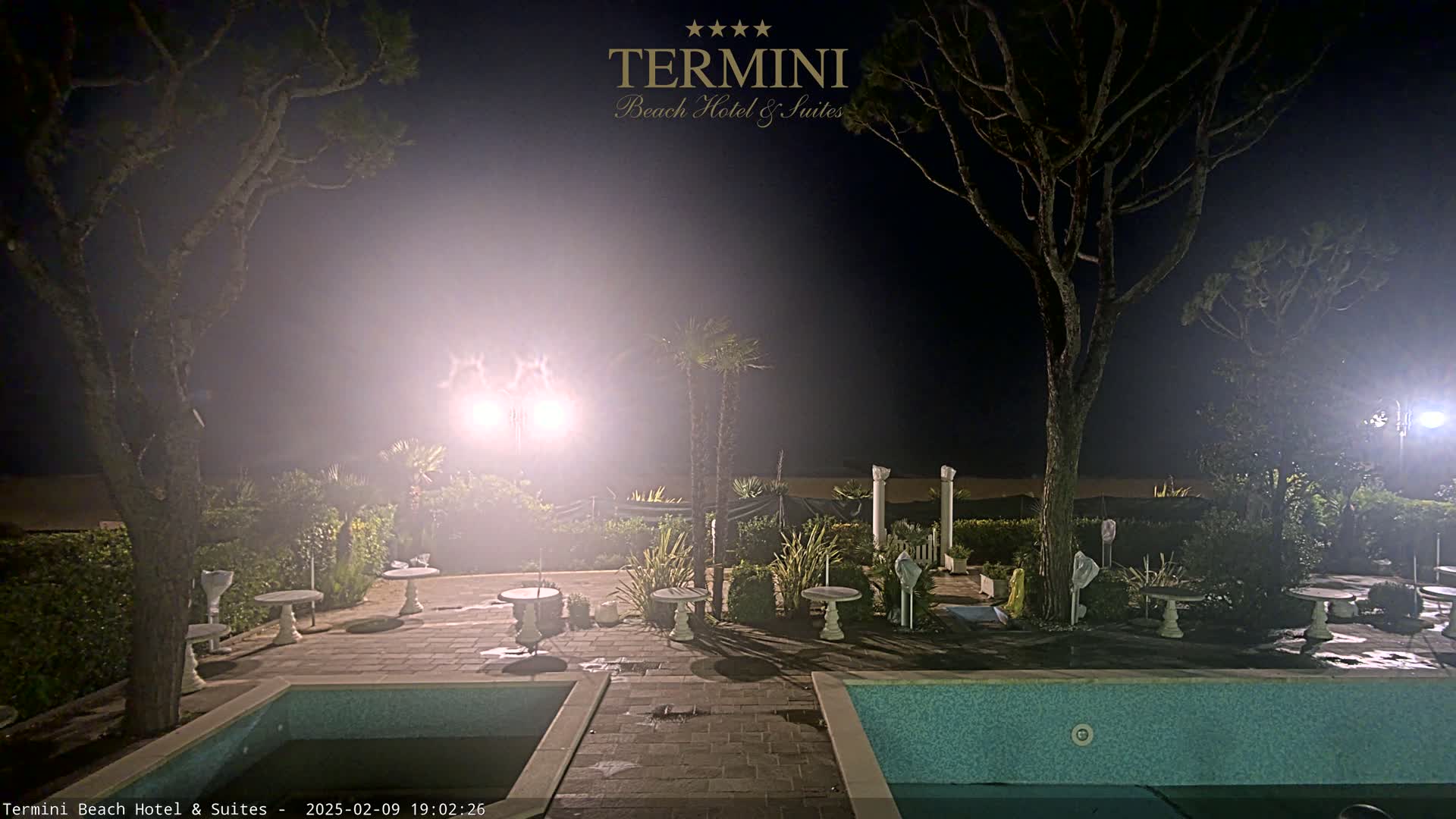 A hazy night scene shows a patio area with a swimming pool, small tables, and trees surrounding it, illuminated by bright lights.