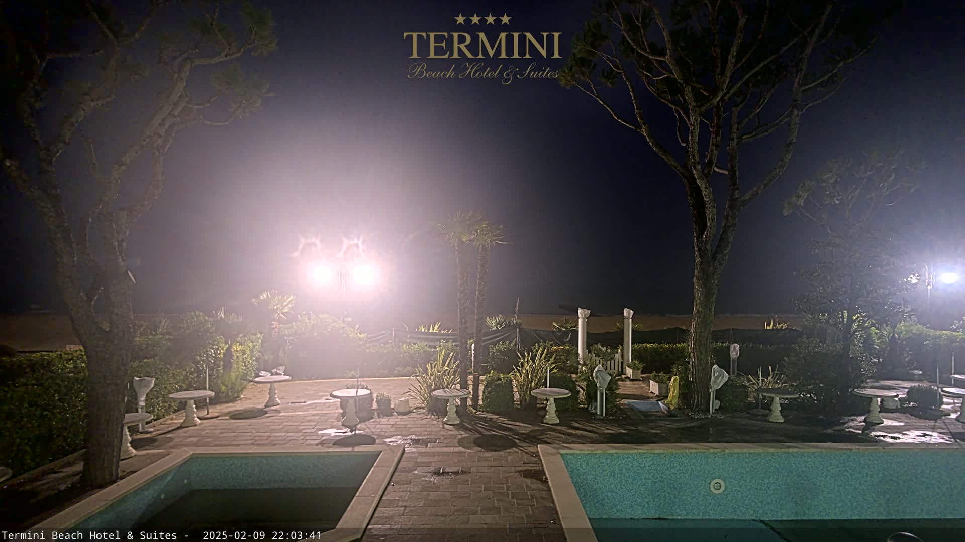 A hazy night scene shows a poolside patio area with small tables and trees, illuminated by bright lights, overlooking a beach.