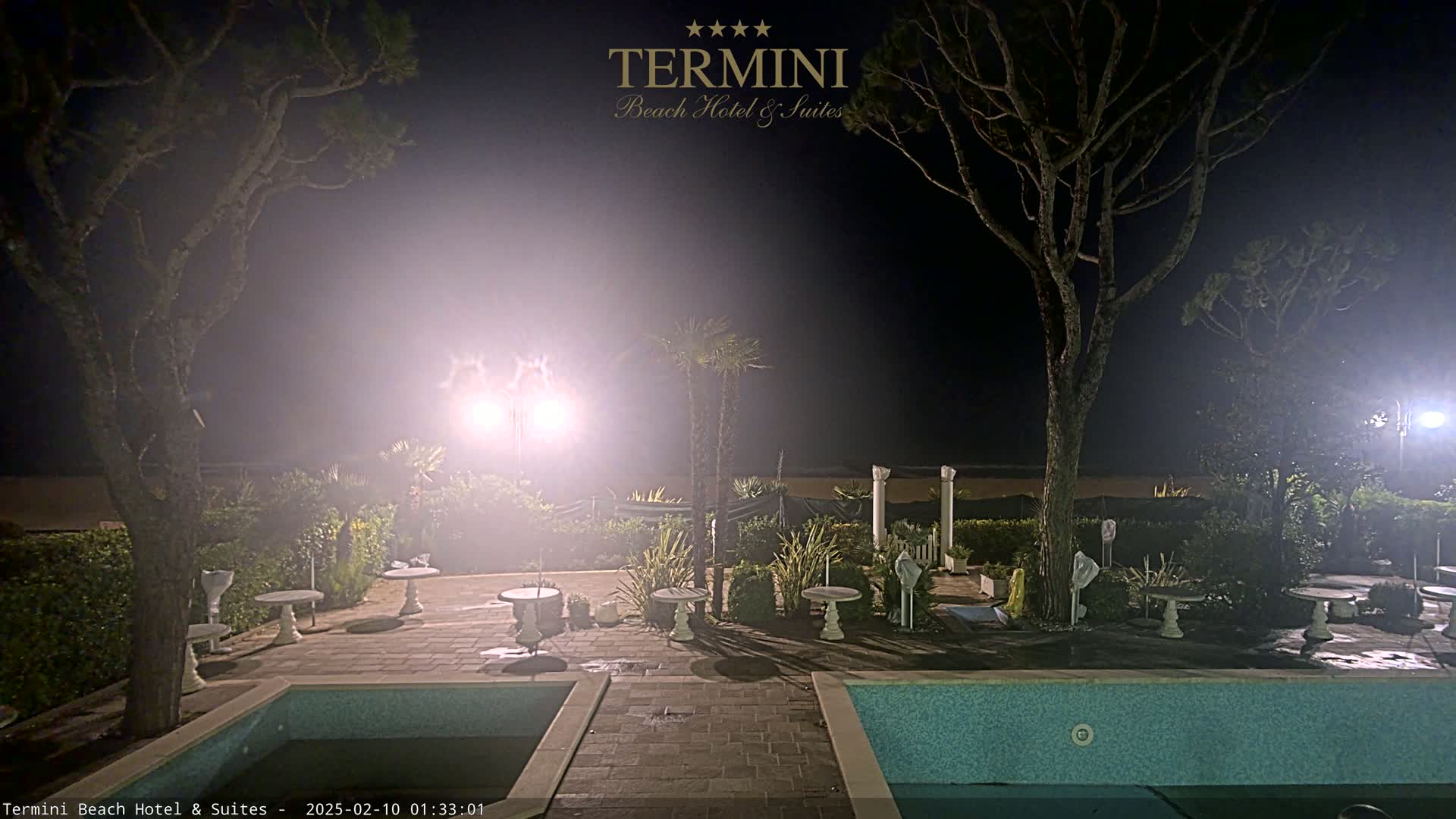 A hazy night scene shows an outdoor patio area with small tables, a swimming pool, and trees surrounding the space, illuminated by bright lights.
