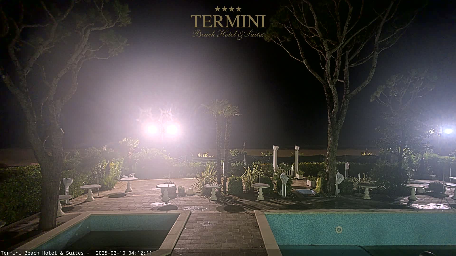A hazy nighttime scene shows a patio area with several small tables and a swimming pool, surrounded by trees and shrubs, with bright lights illuminating the space.