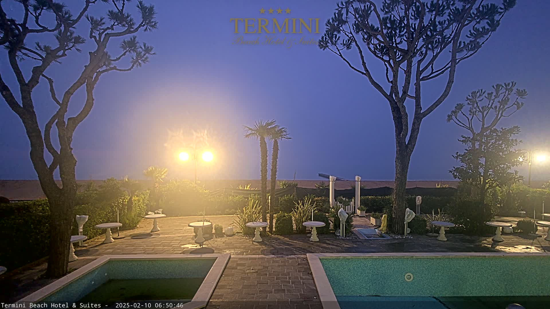 A twilight view of an outdoor patio area with two pools, several small tables, and various trees and plants, overlooking a sandy beach under a clear sky.