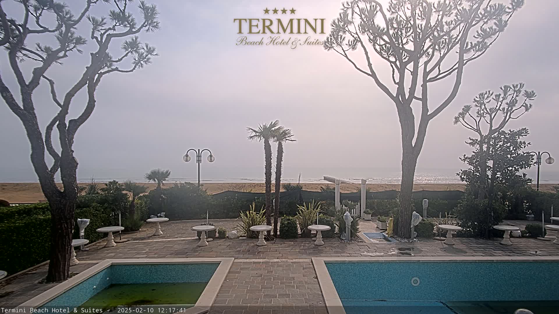 A hazy beach is visible beyond a patio with two swimming pools, several small tables, and various trees and shrubs.