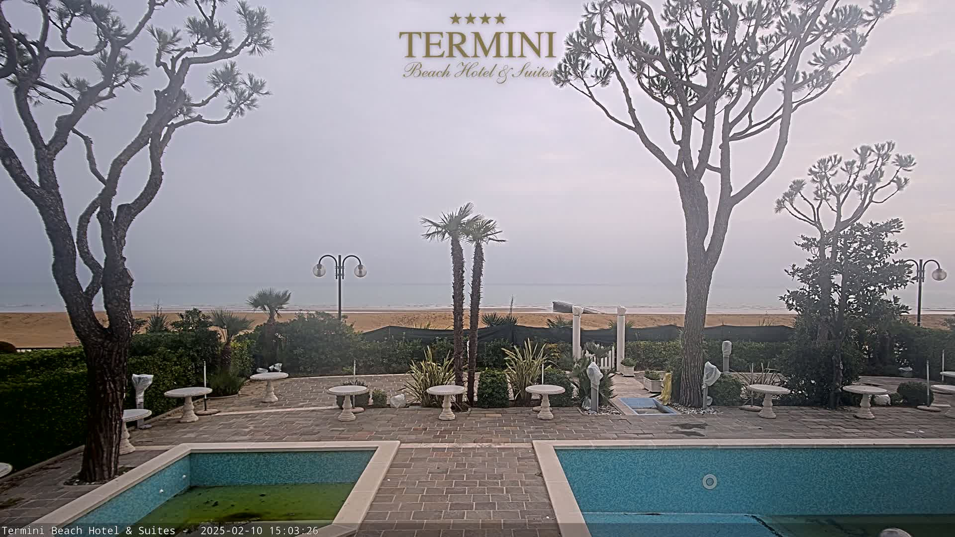 A hazy view of a sandy beach and calm ocean is seen from a patio area with two swimming pools, some palm trees, and several small tables.