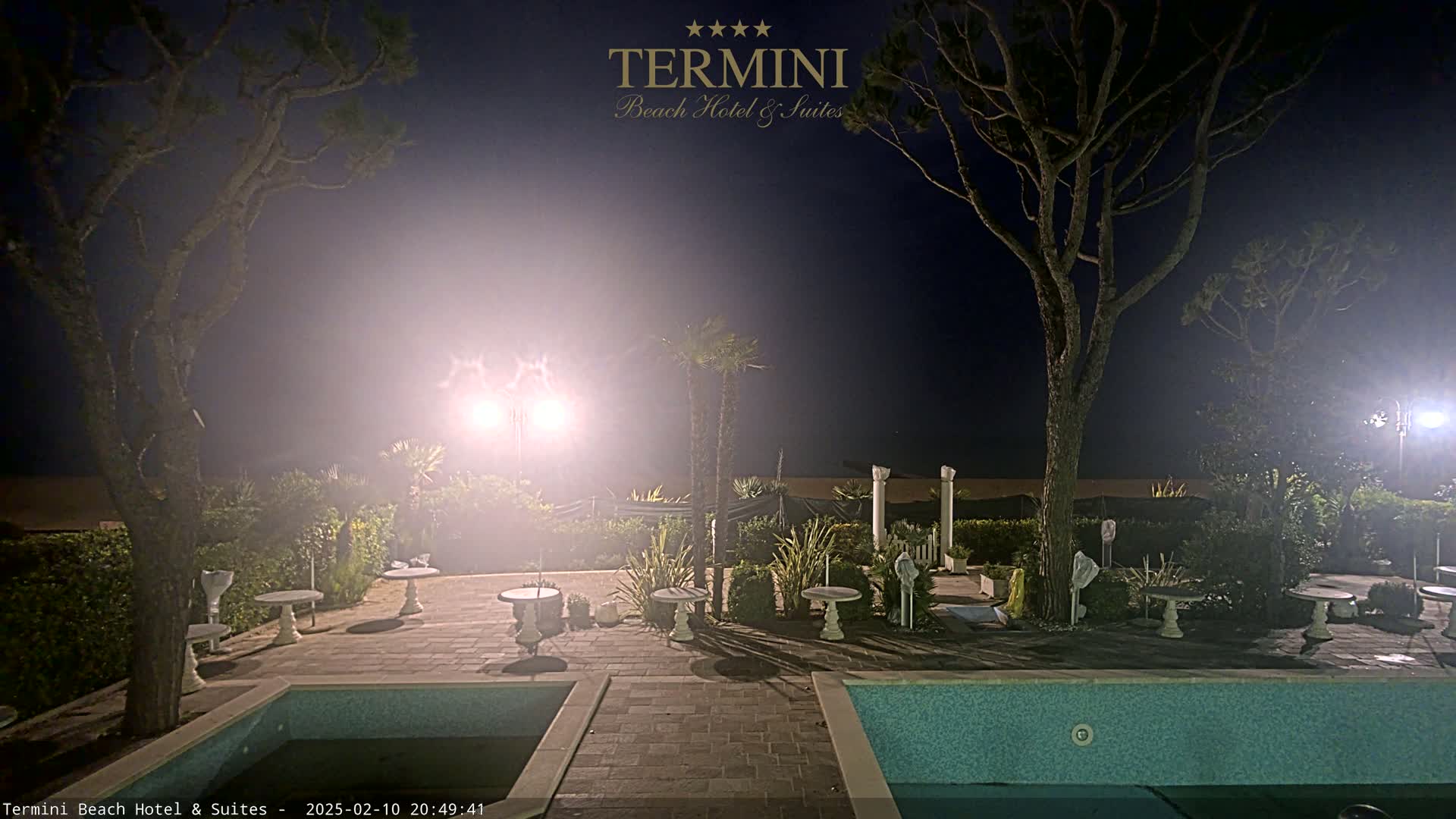 A nighttime outdoor scene shows a pool and patio area with small tables and trees, illuminated by bright lights under a hazy, dark sky.