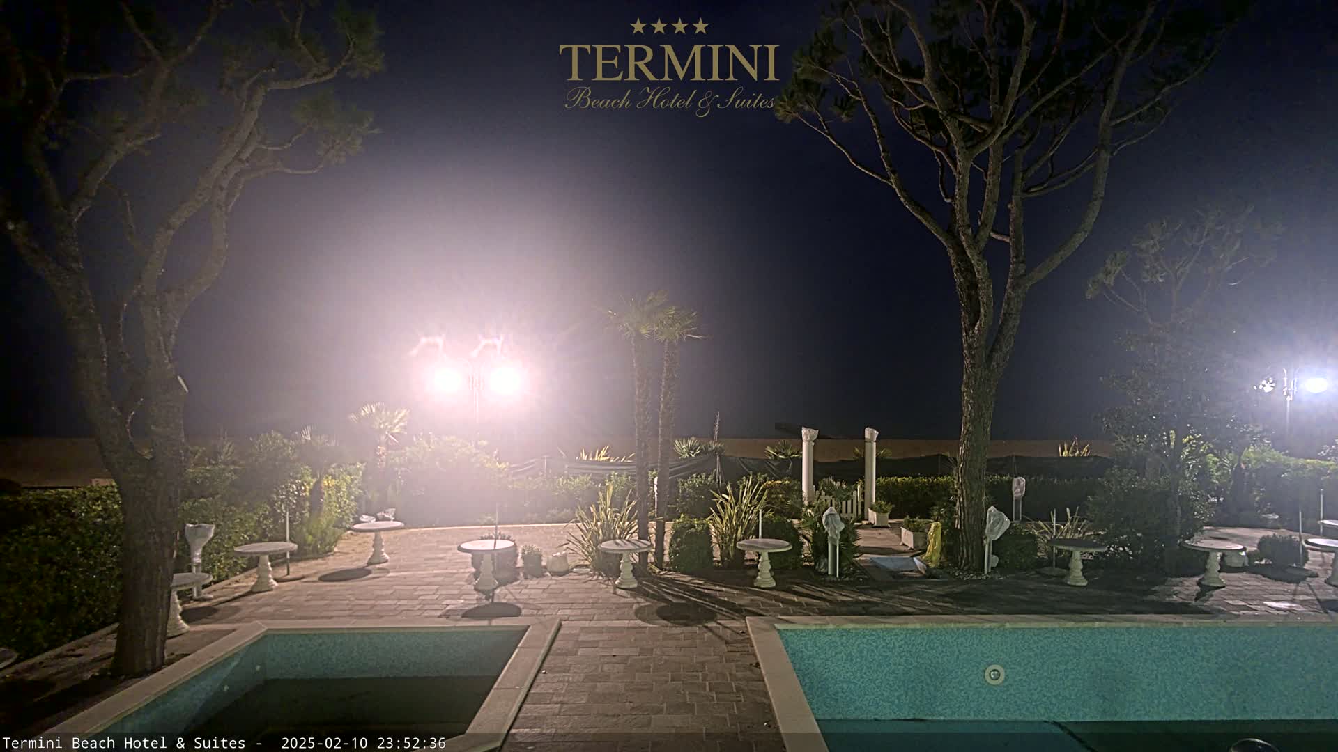 A nighttime view of a patio area with a swimming pool, small tables, and trees under hazy, bright lights.