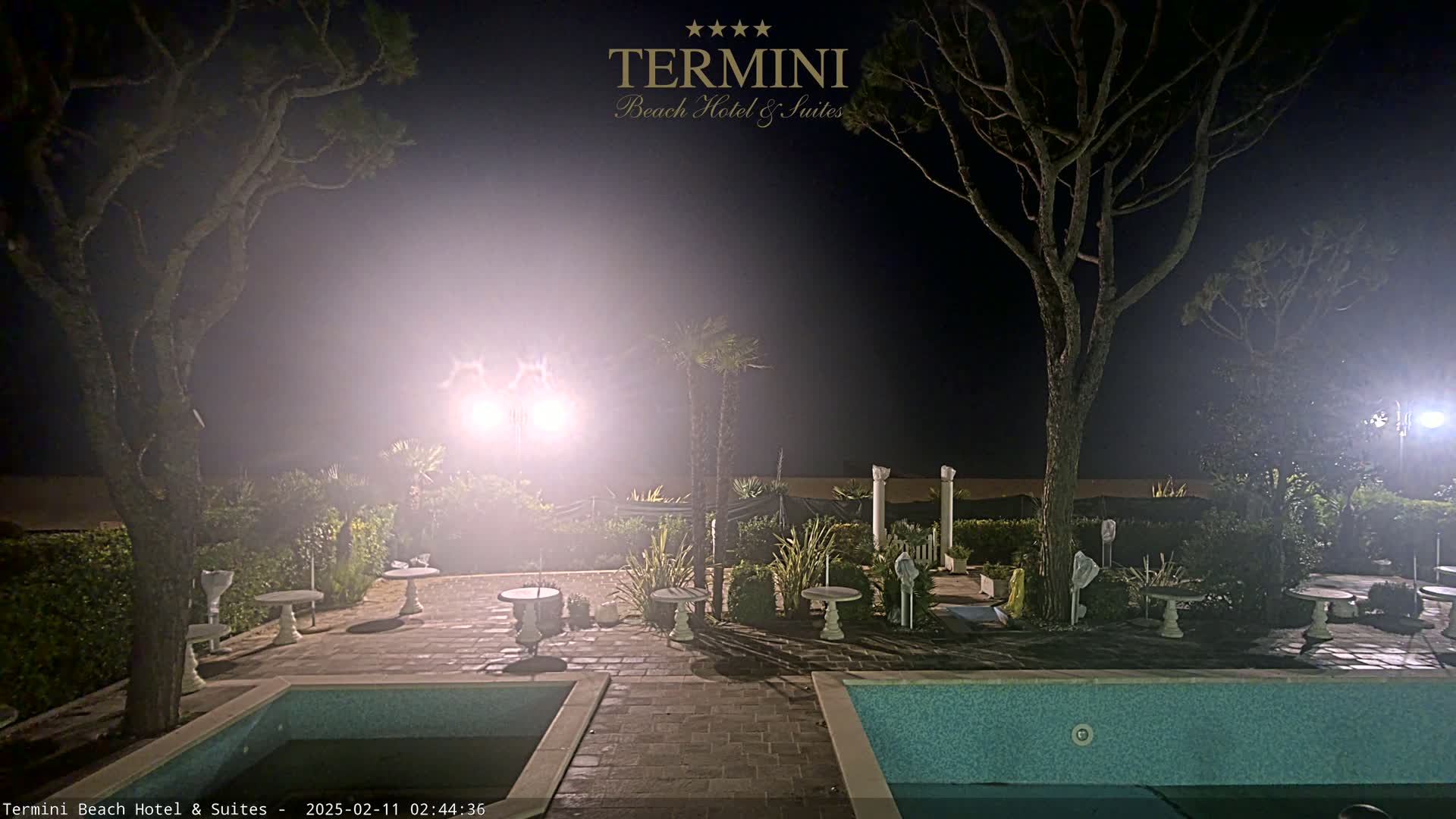 A hazy night scene shows a patio area surrounding a swimming pool, lit by bright lights, with trees and small tables scattered around.